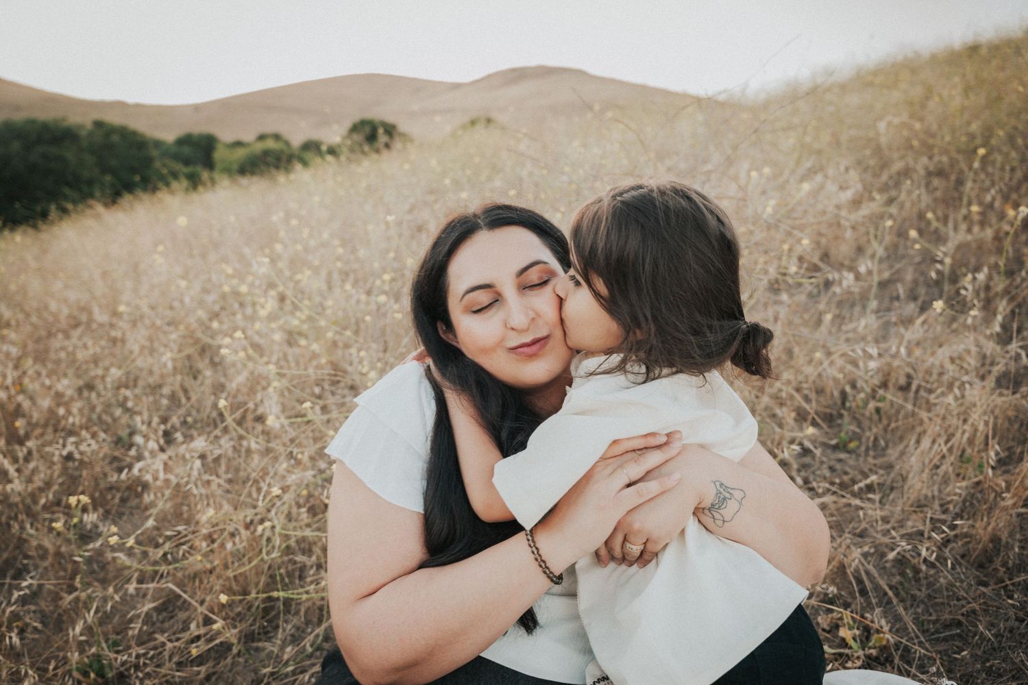 A mother smiles warmly as her young child gives her a tender kiss on the cheek in a golden field.