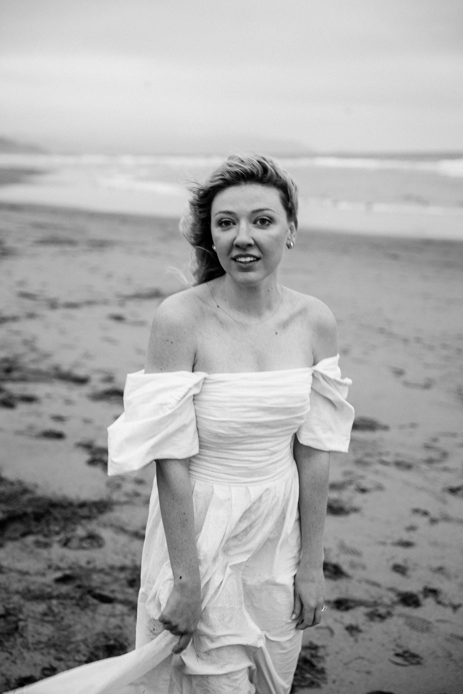 Bride in elegant off-shoulder white dress standing on a sandy beach with ocean waves in black and white photo.