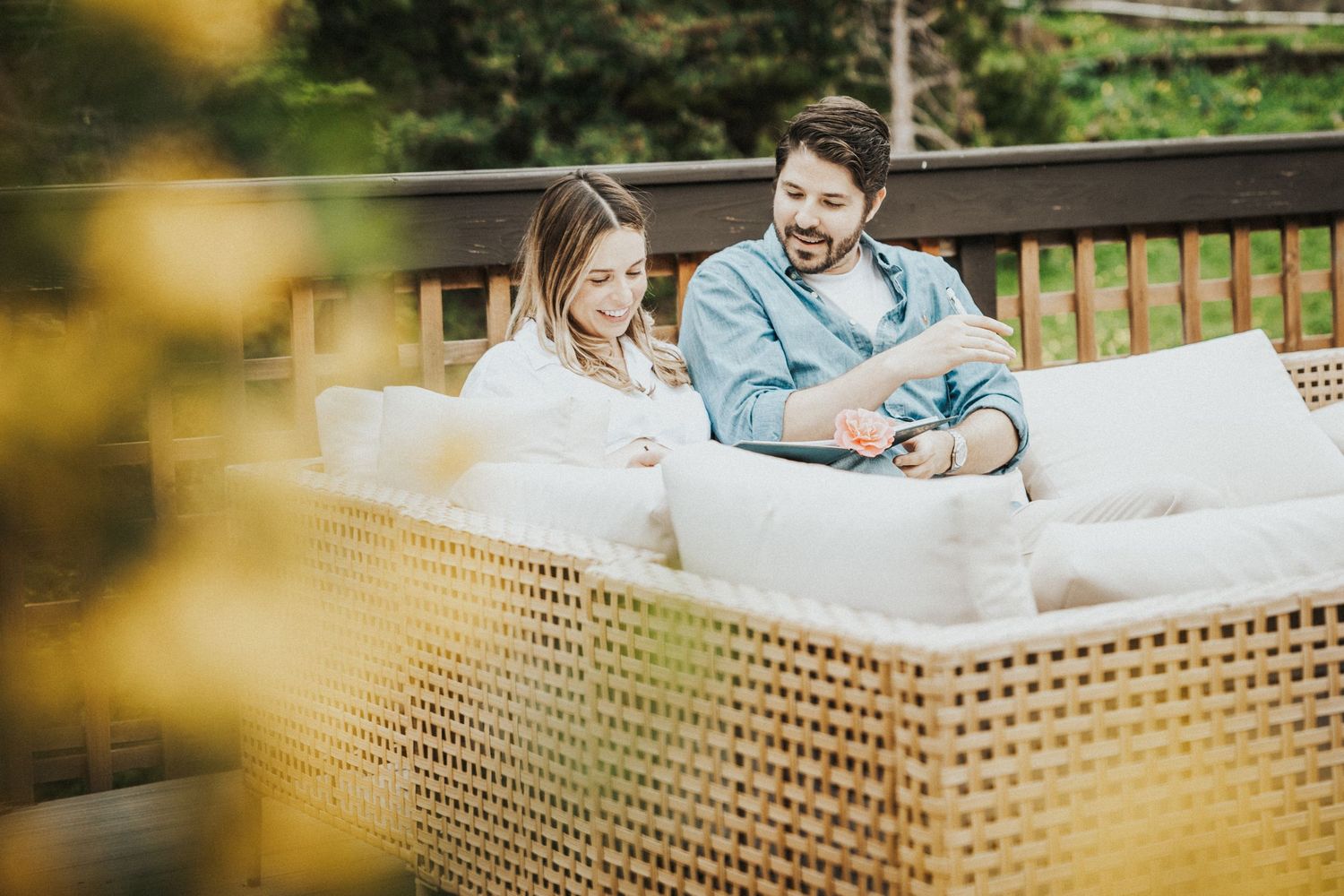 Couple relaxing on outdoor wicker sofa with white cushions on a deck surrounded by greenery.