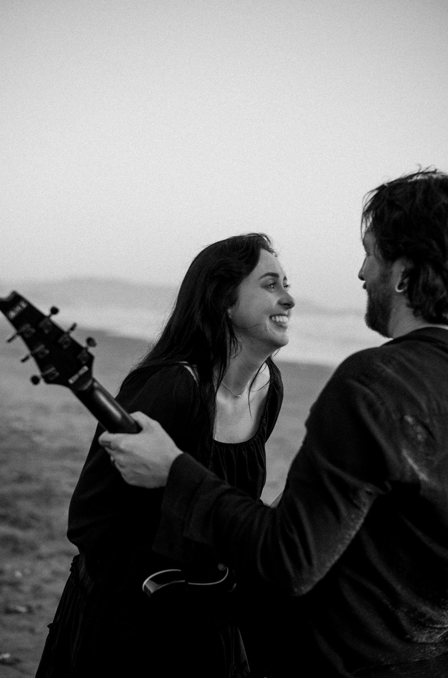Black and white photo of a smiling woman holding a guitar facing a man on a misty beach.