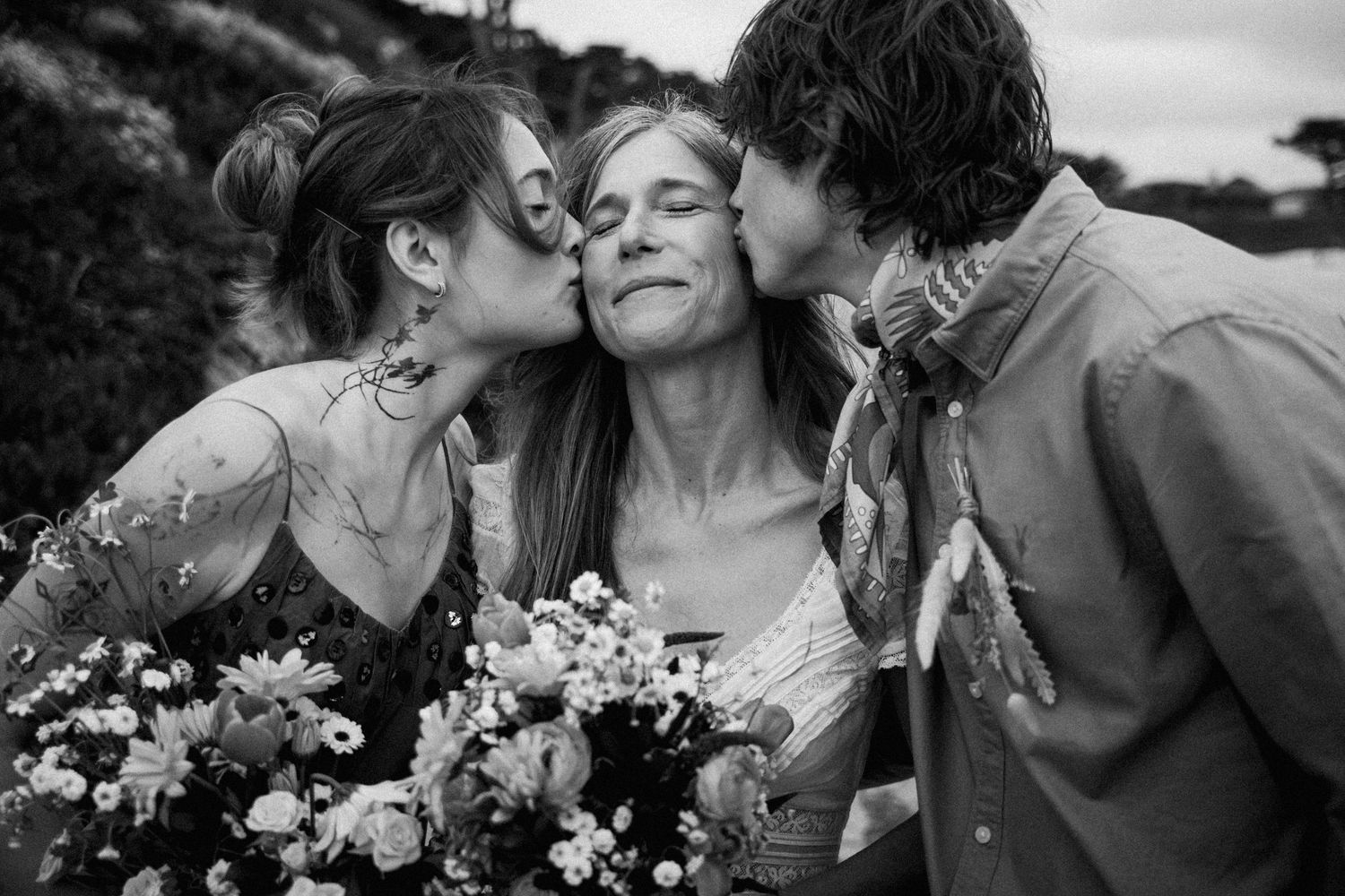 Black and white photo of two people kissing the cheeks of a smiling woman holding a wildflower bouquet outdoors.