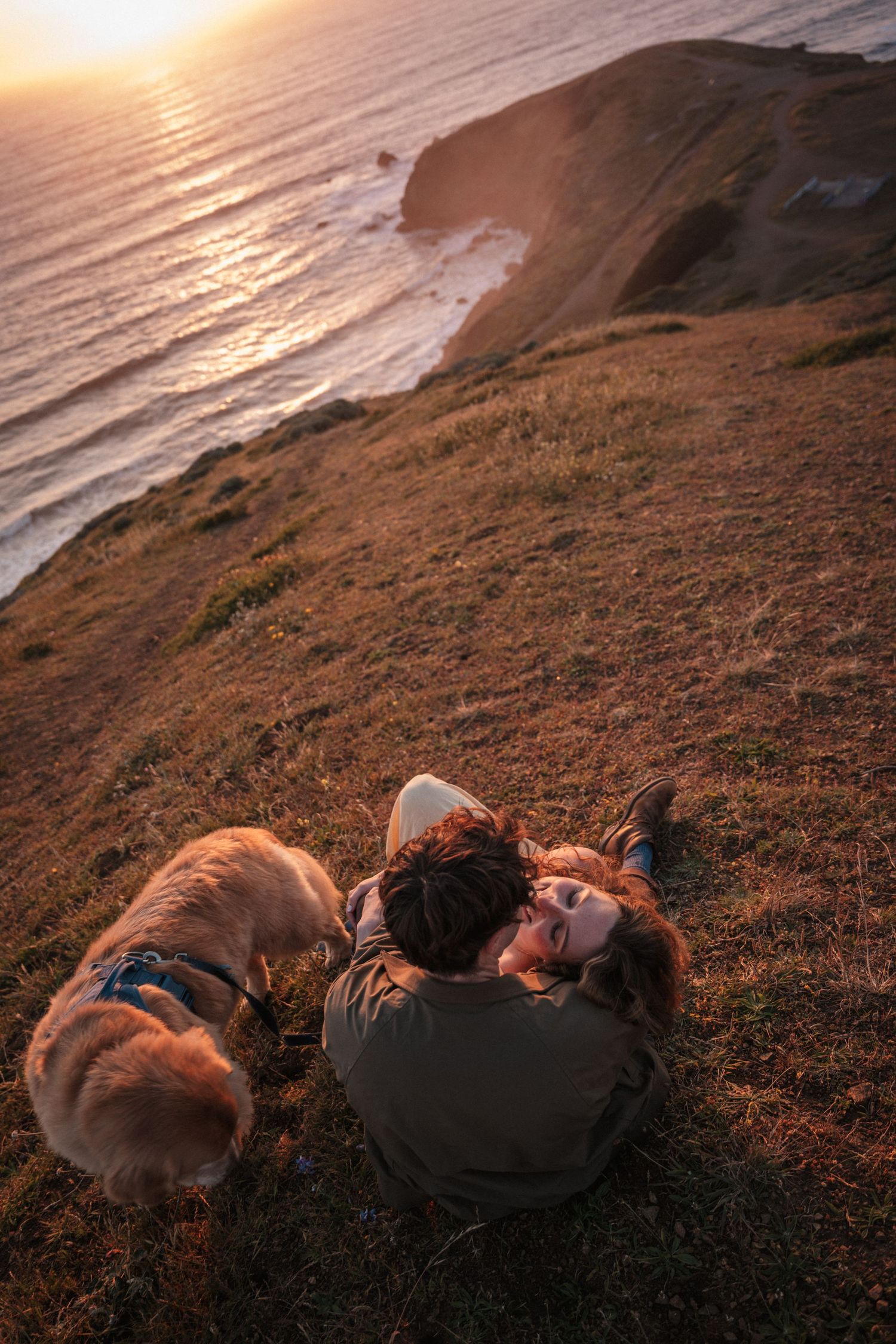 Aerial view of couple lying on grassy cliff with dog enjoying golden sunset over ocean.