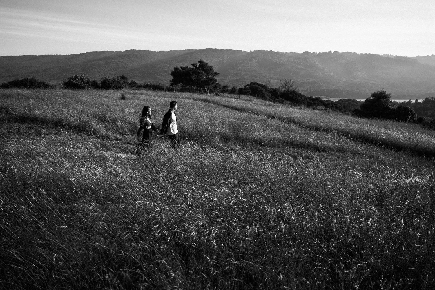 Two people walking through tall grass on a scenic hillside in black and white photography.Photography by The Y.K. Studio.