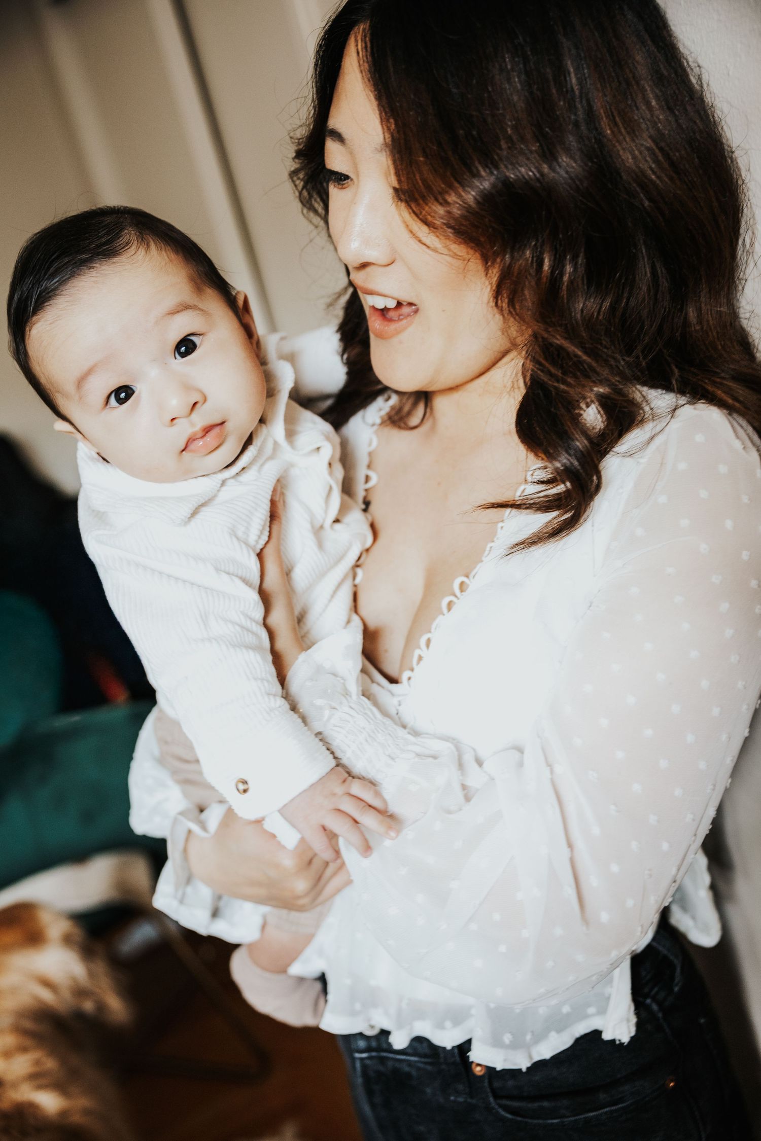 A woman in a white lace blouse tenderly holds her infant baby, smiling warmly in a cozy home setting.