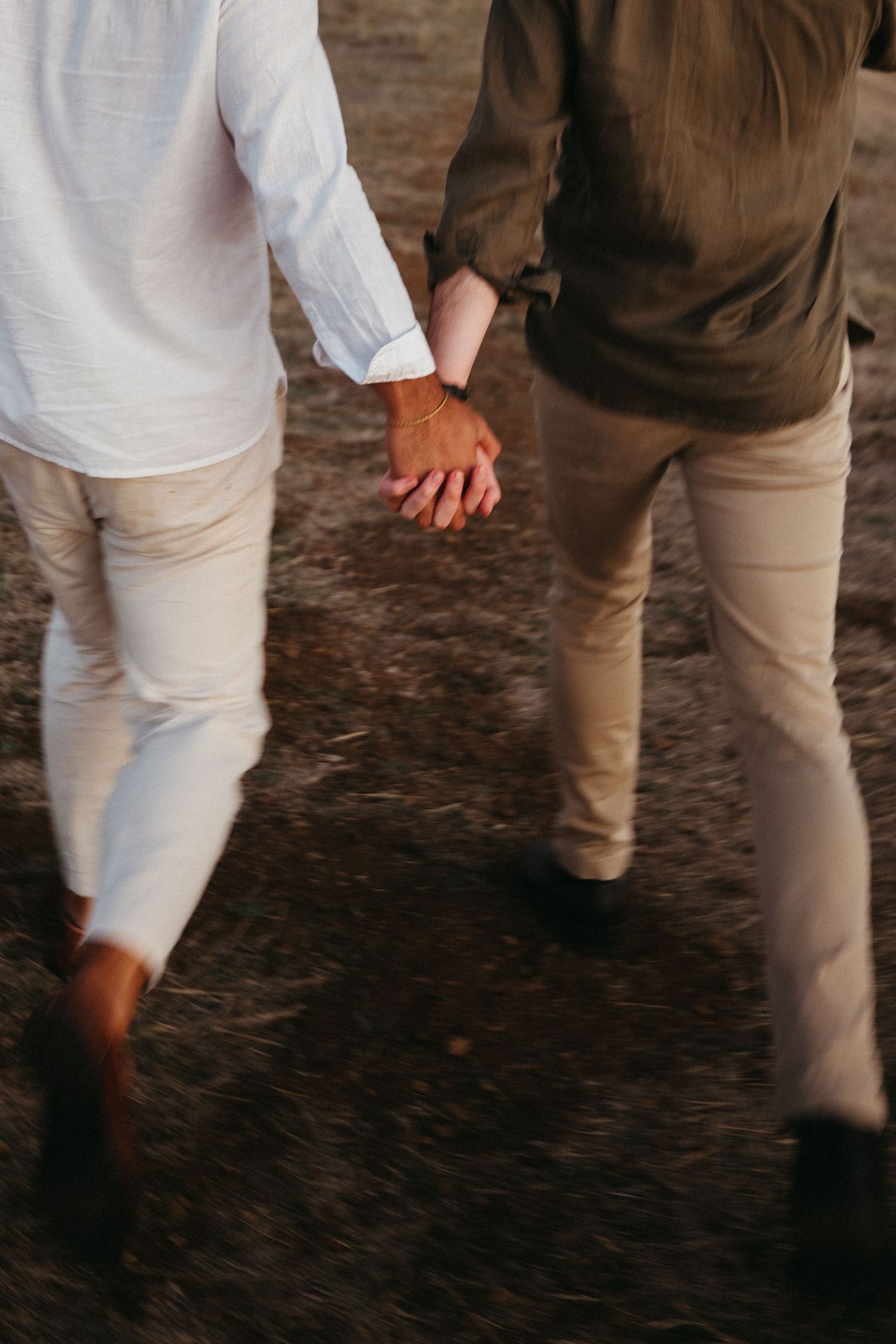 Couple holding hands while walking outdoors, one in white shirt and beige pants, other in olive jacket and khaki trousers.