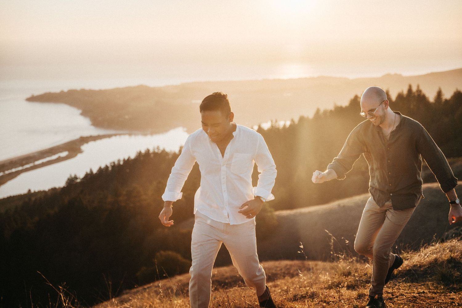 Two men hiking on a mountain trail at golden sunset with scenic river valley views in the background.