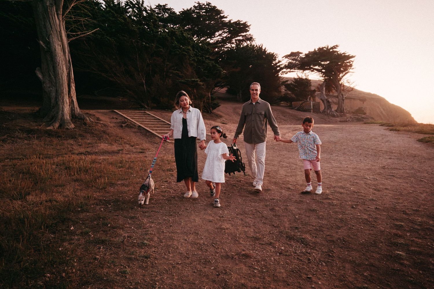 Family of four with a dog walking on a scenic coastal trail at sunset with lush green trees in the background.