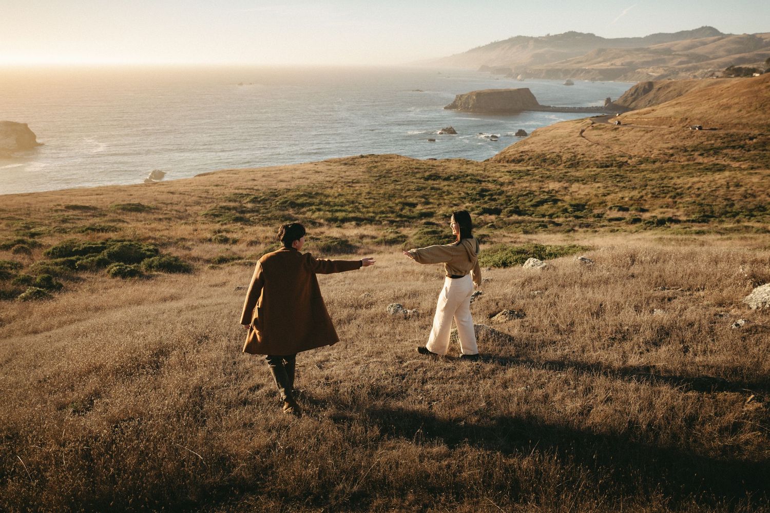 Two people walk hand in hand across a golden grassy hillside overlooking the ocean and rugged coastline at sunset.