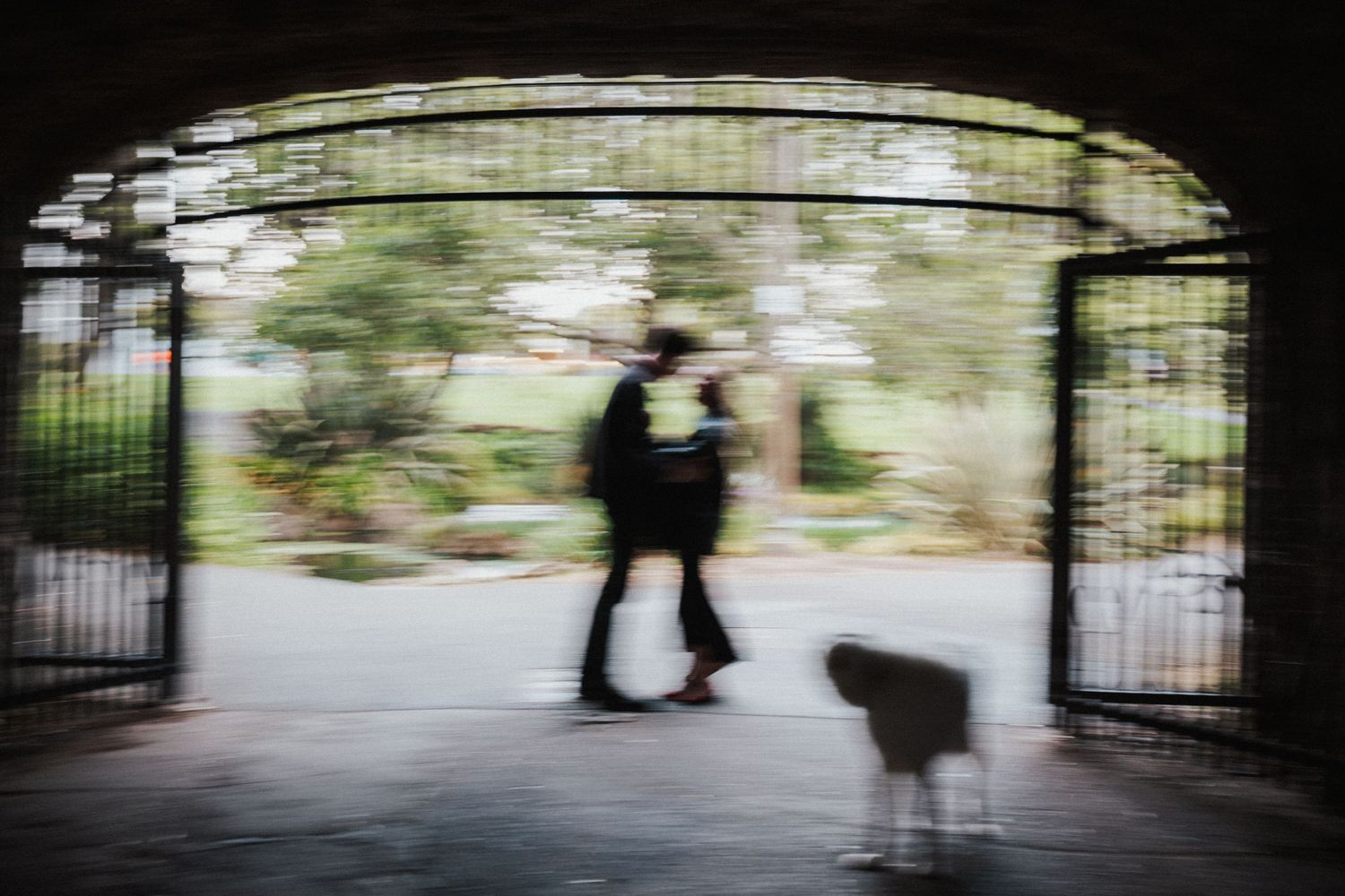 Blurred silhouettes of a couple walking a dog through a stone archway in a scenic park.Photography by The Y.K. Studio.