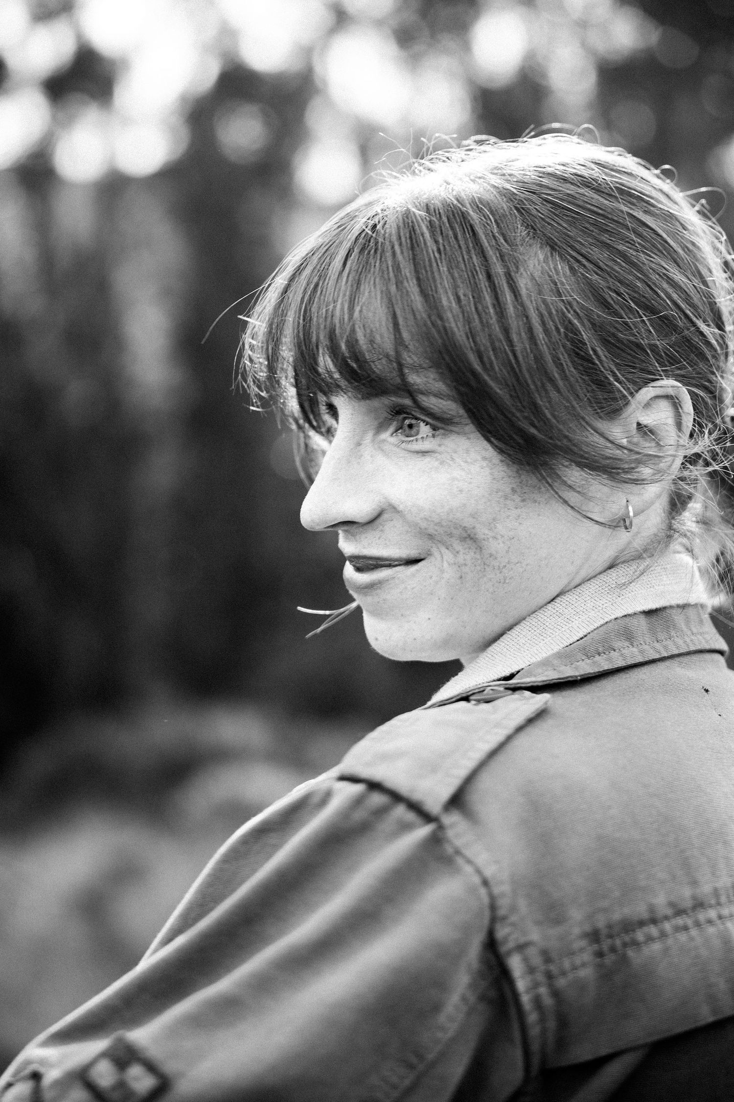 Black and white portrait of a smiling woman with bangs and freckles, looking over her shoulder outdoors.Photography by The Y.K. Studio.