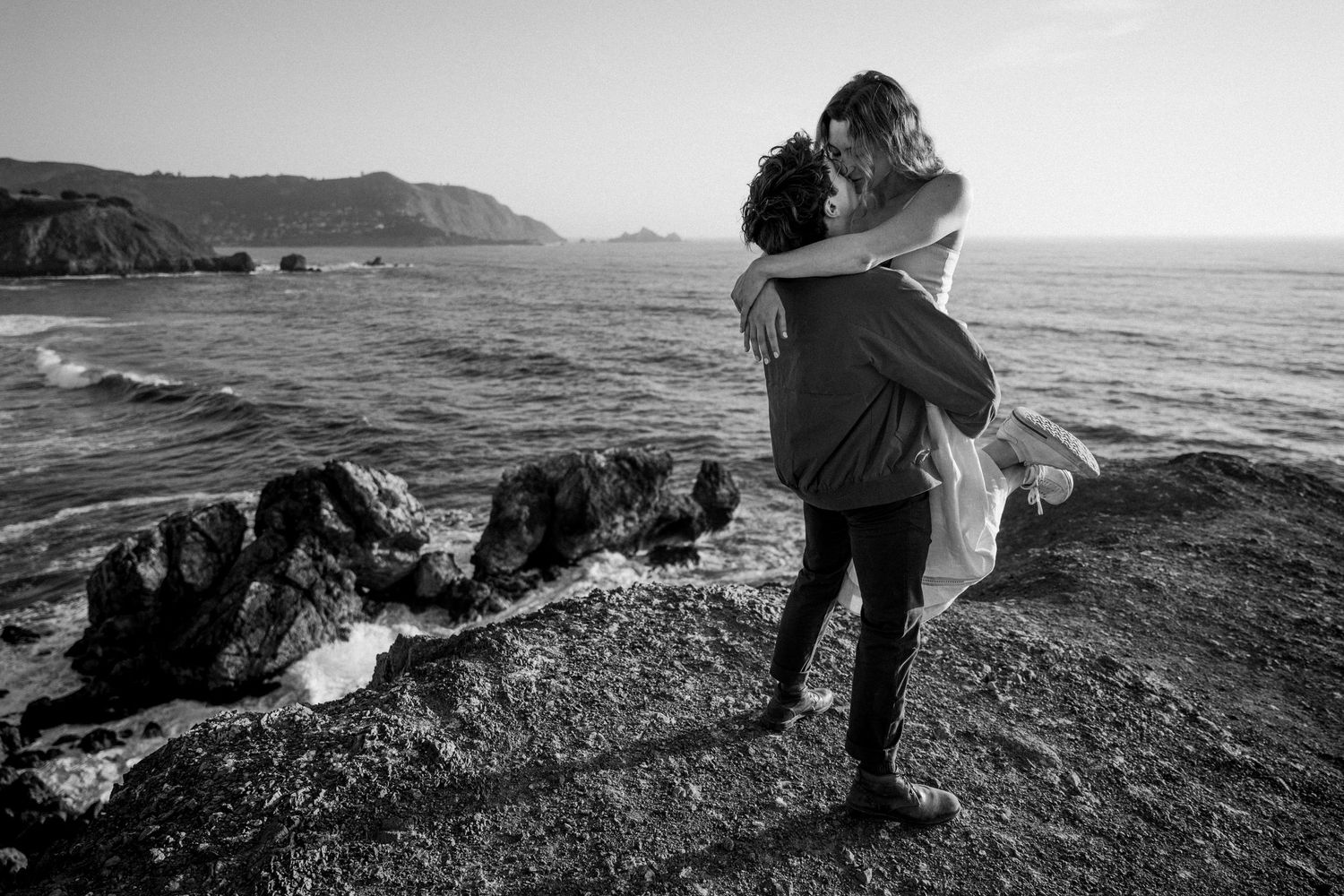 Couple embracing on rocky coastline in black and white, man lifting woman with ocean and cliffs behind them.