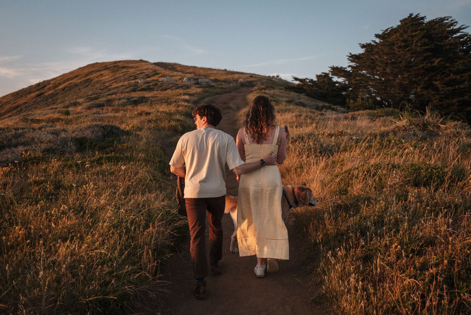 Couple walking hand in hand along a golden hillside trail at sunset.