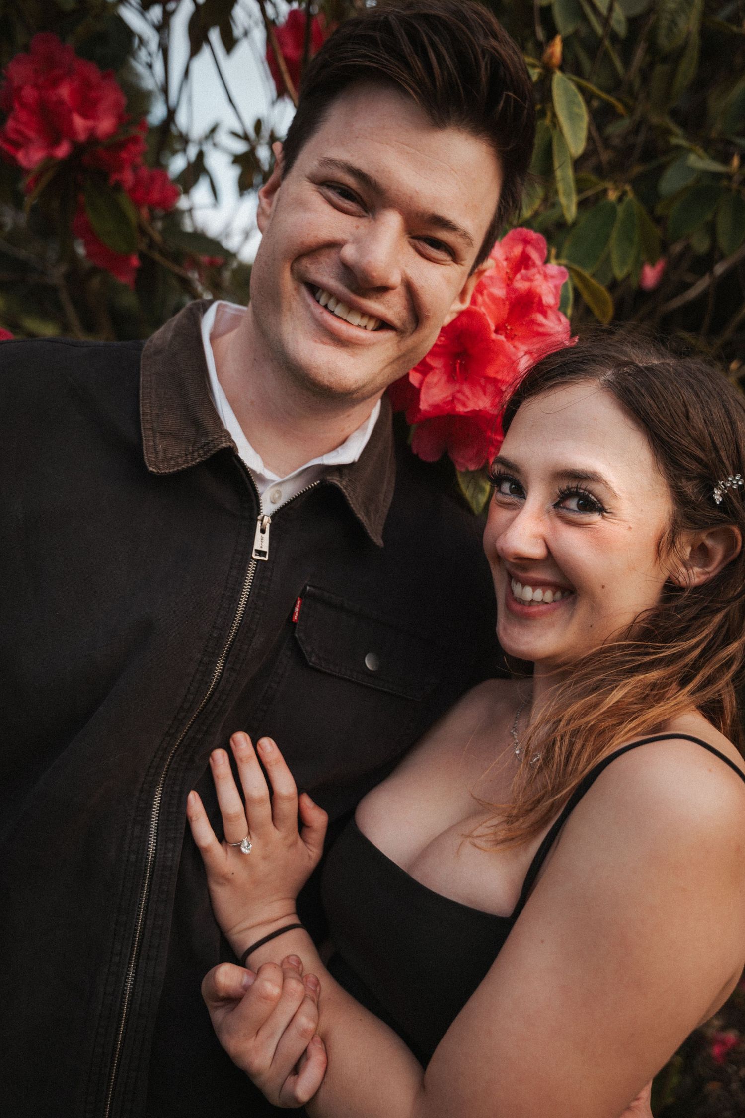 Smiling engaged couple embracing in front of vibrant red rhododendron flowers outdoors.