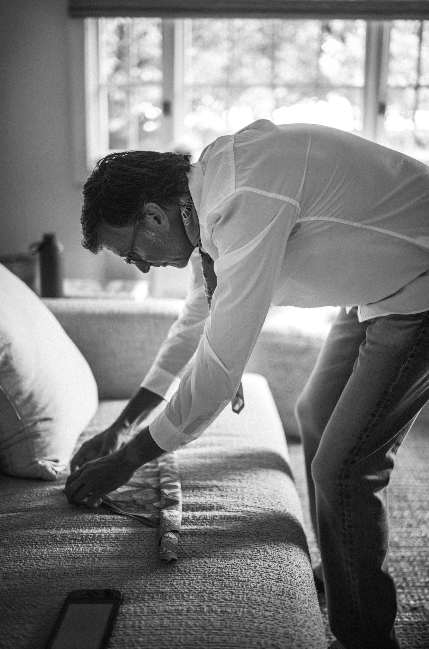 Man in white shirt leaning over sofa arranging cushions in black and white photo.