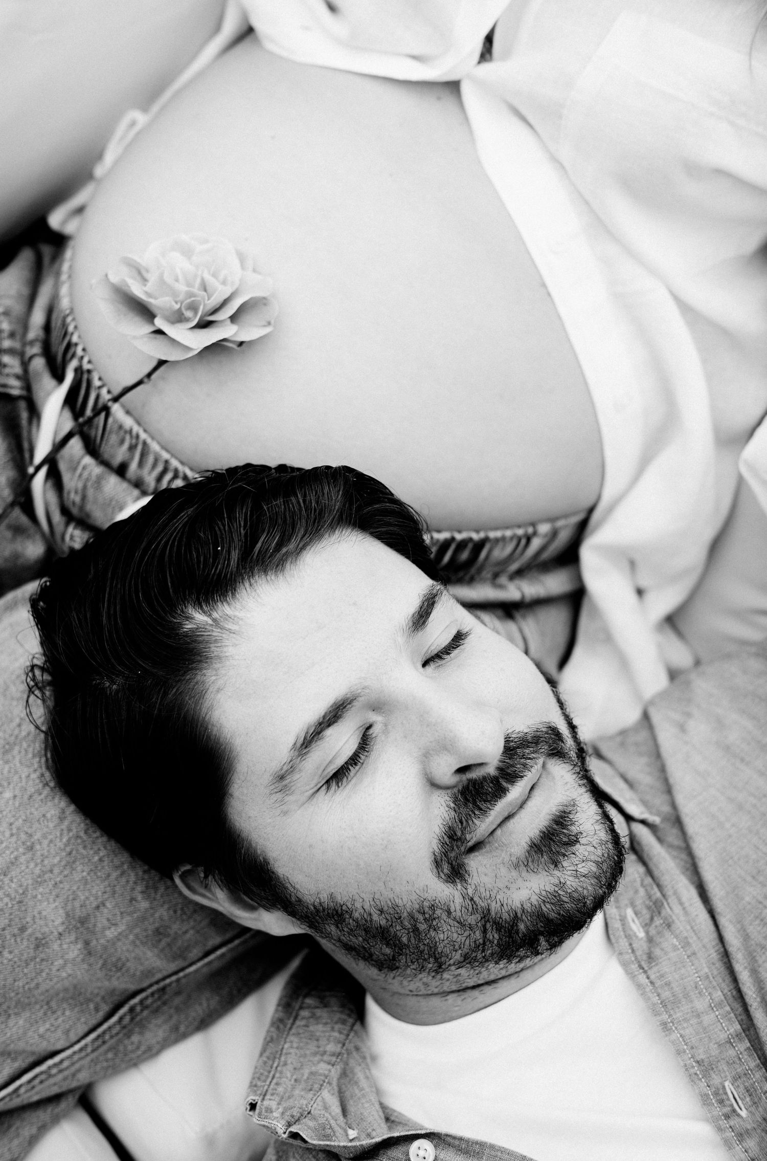 Black and white maternity photo of a man resting his head on a pregnant womans belly with a flower decoration.