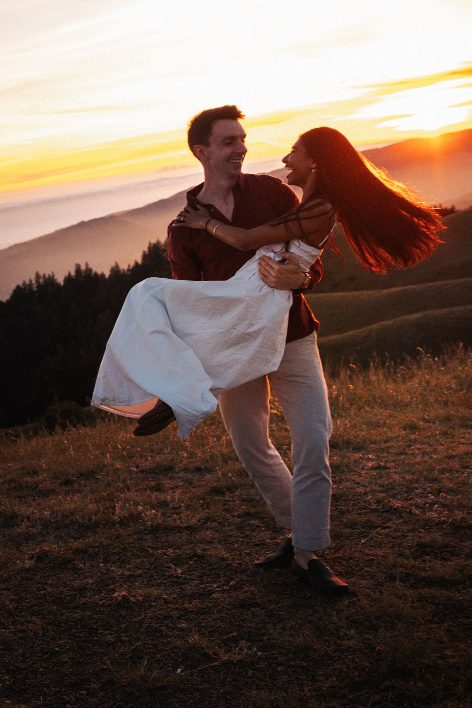 A joyful couple embraces at sunset on a mountain meadow, man lifting woman in white dress against golden sky.