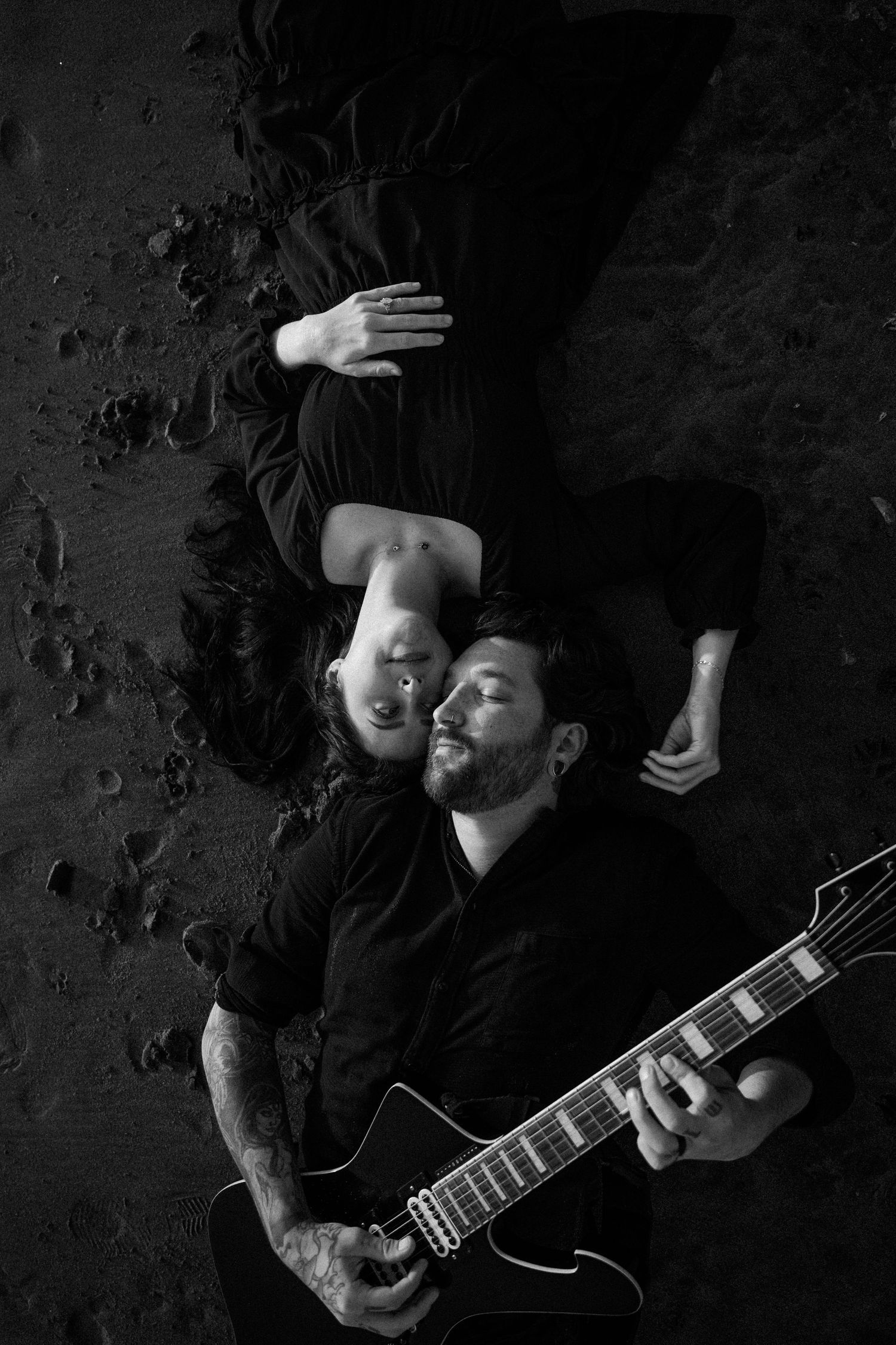 Man with beard plays electric guitar lying on dark sand beside a woman in black and white photo.