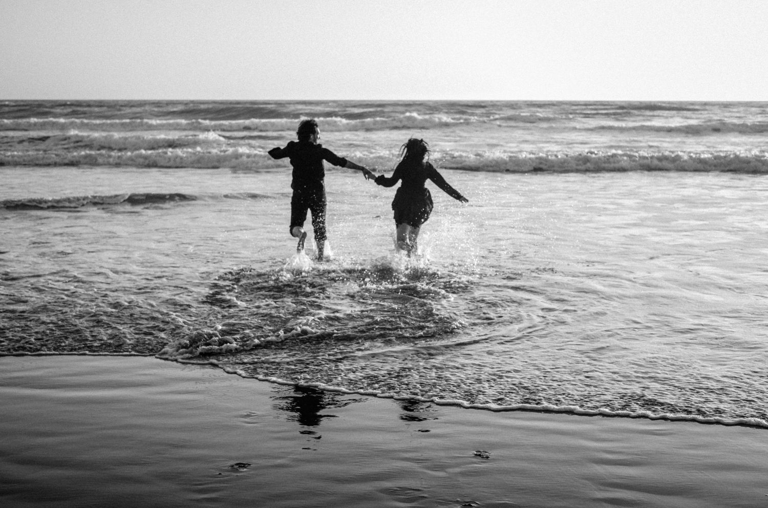 Two children holding hands and splashing in ocean waves on a beach in a black and white photo.