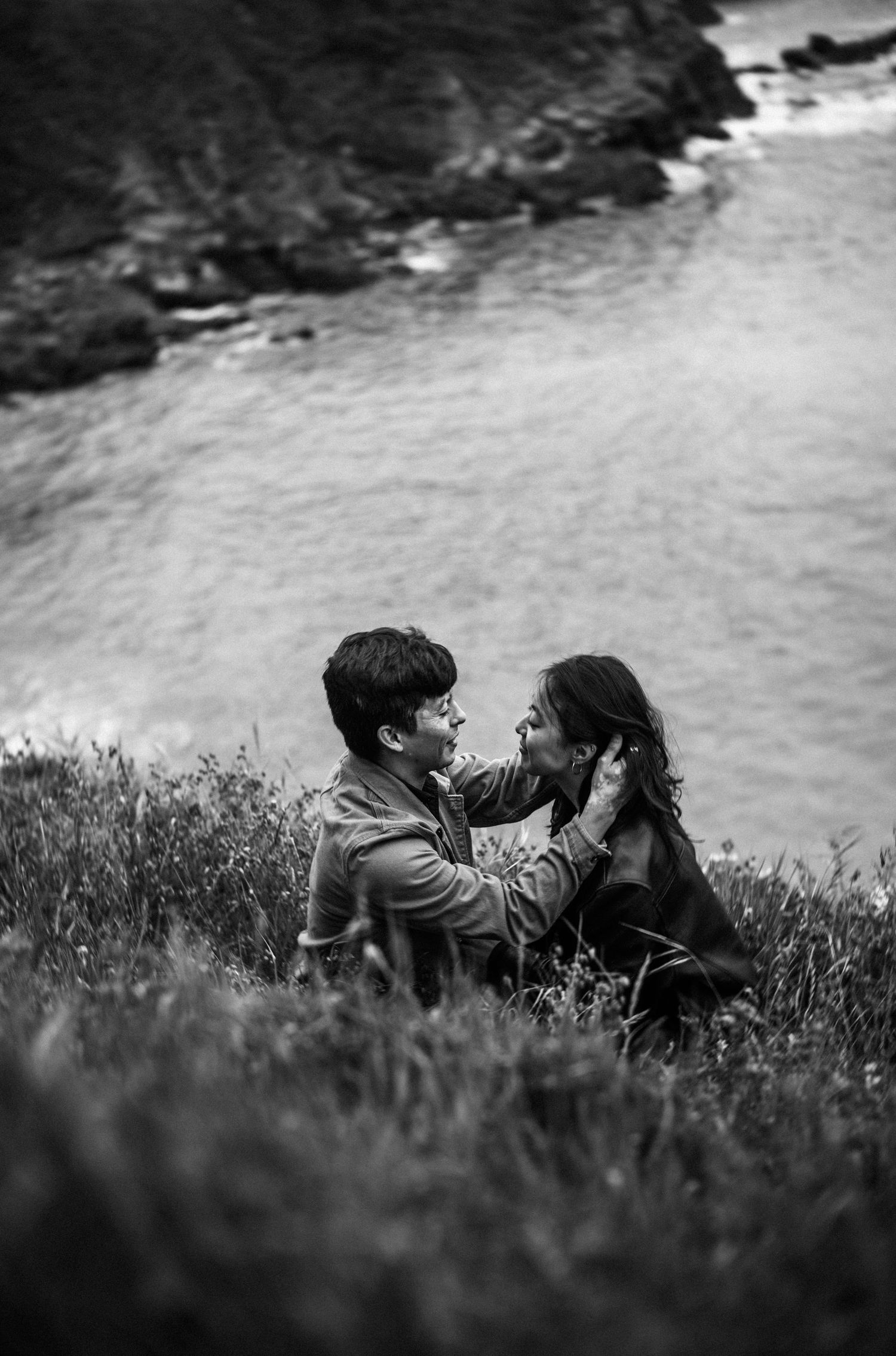 Black and white photo of couple sitting on coastal cliffside grass with ocean backdrop.