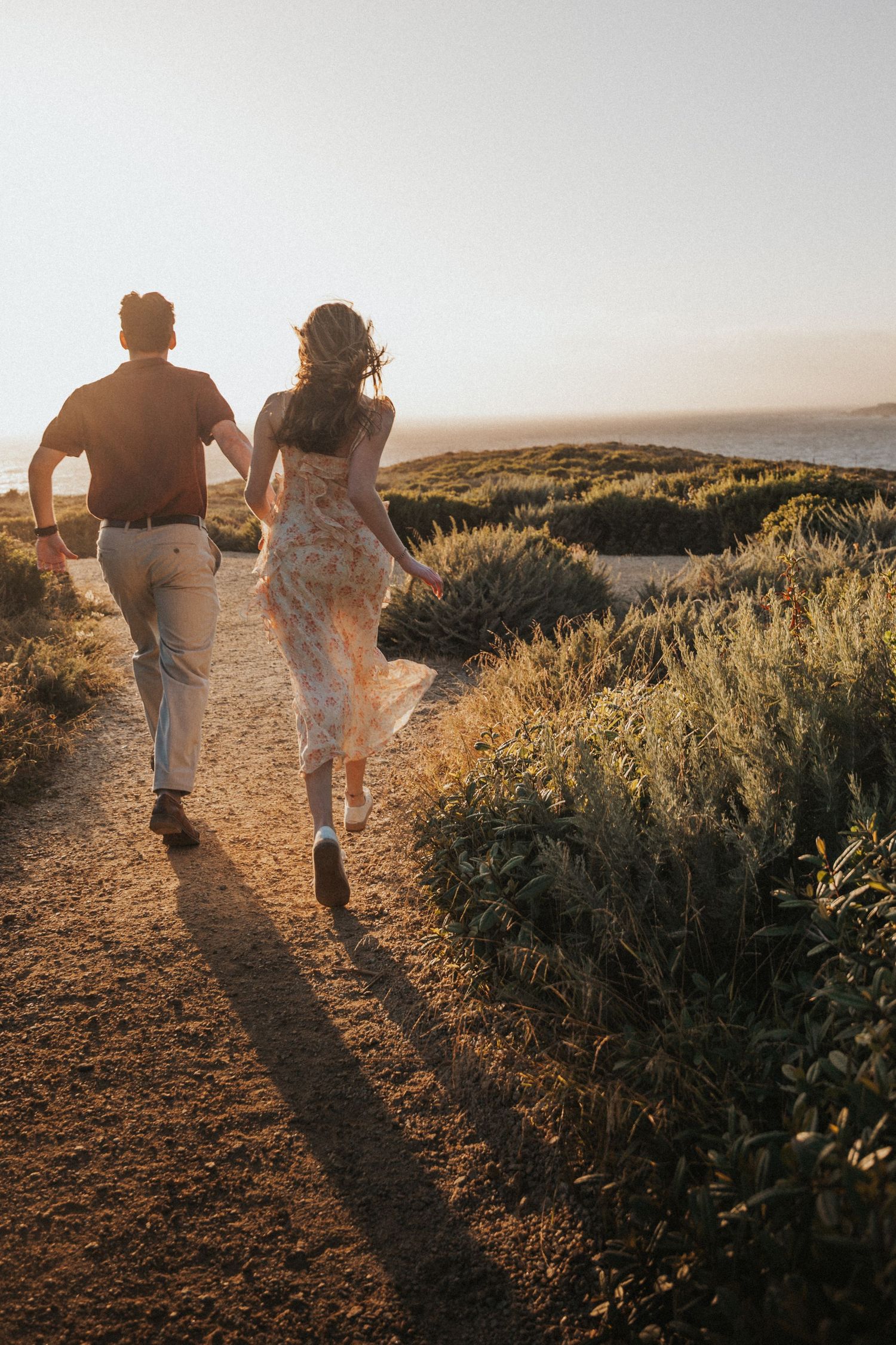 Couple holding hands running along a scenic coastal path bathed in warm golden sunset light.