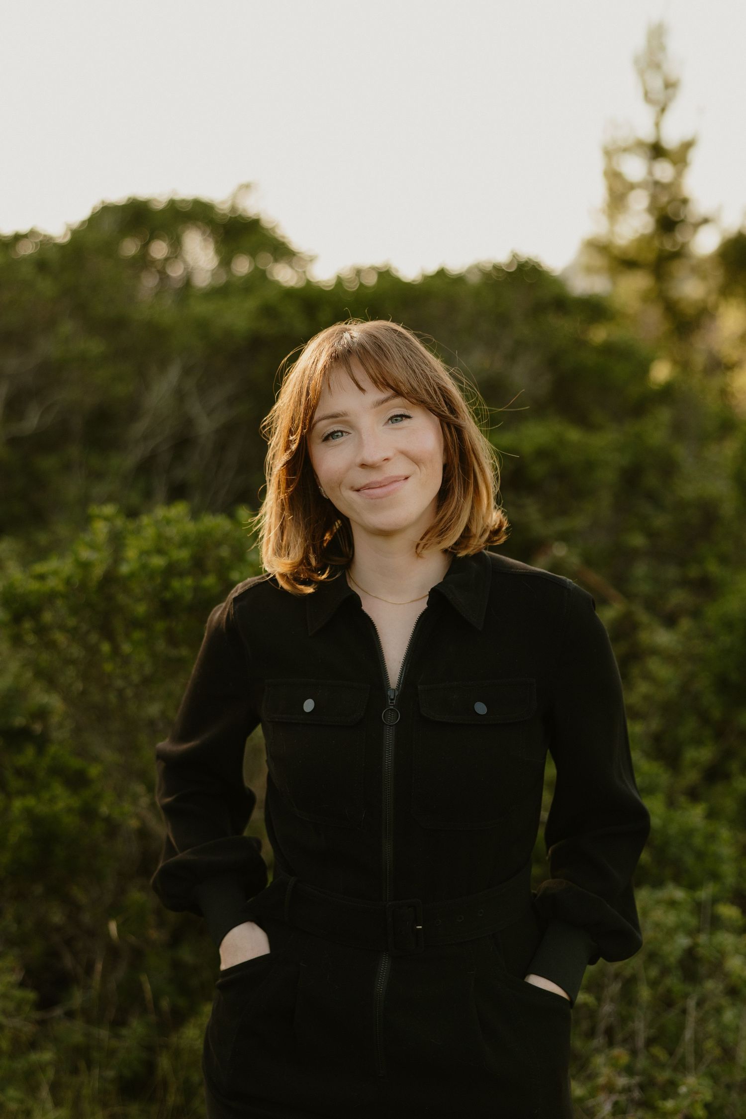 Young woman with short auburn hair smiles outdoors in black jumpsuit with green trees in background.