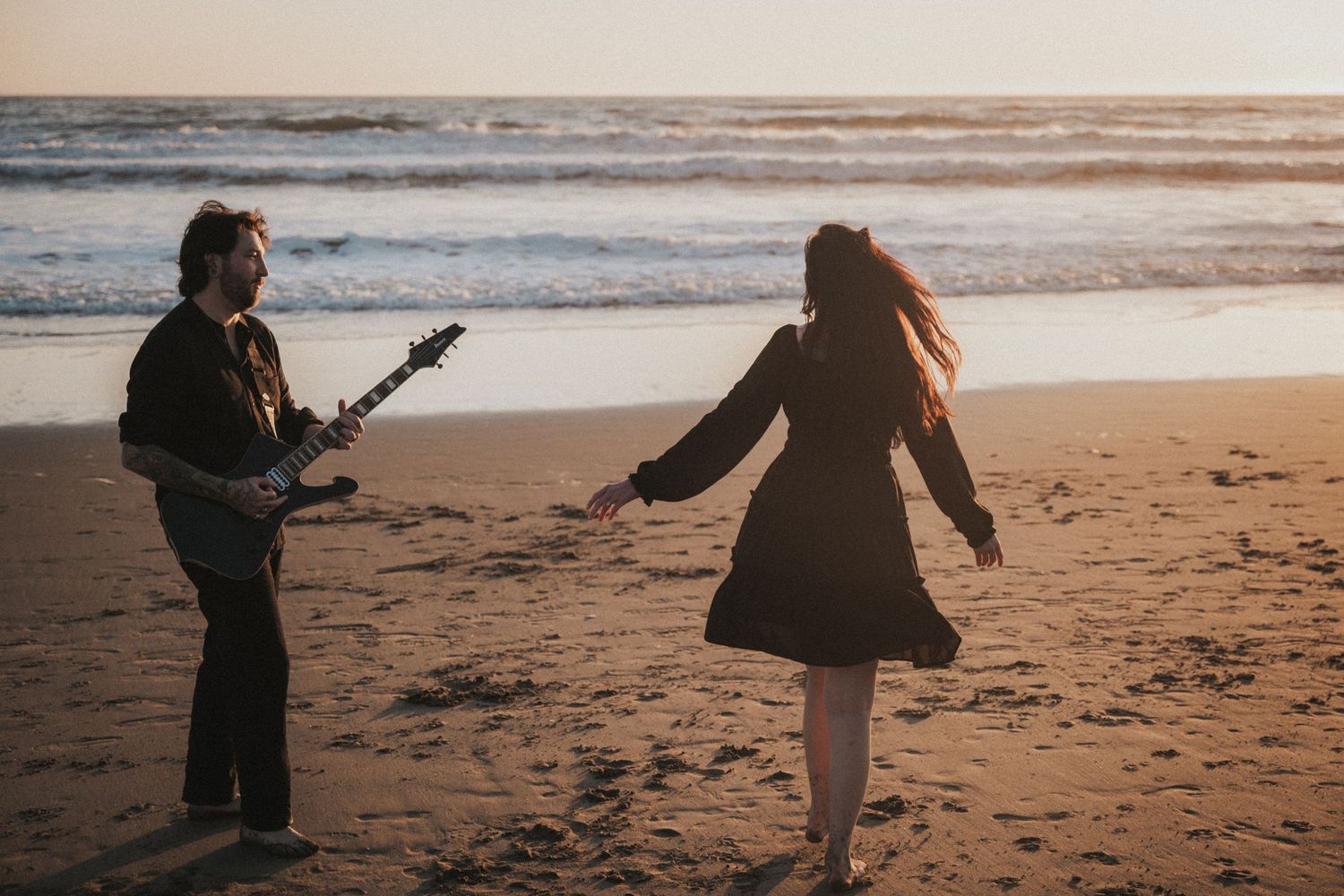 Man playing electric guitar and woman dancing on a sandy beach at sunset near ocean waves.