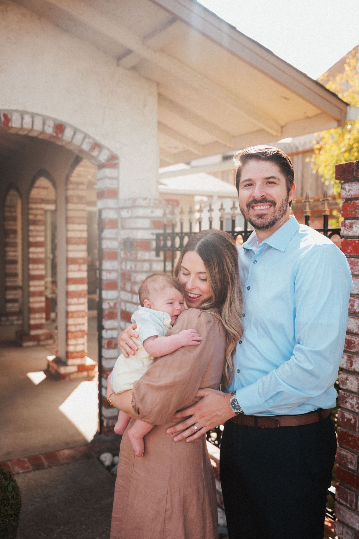Happy young couple smiling outdoors holding their newborn baby near a brick home in warm sunlight.