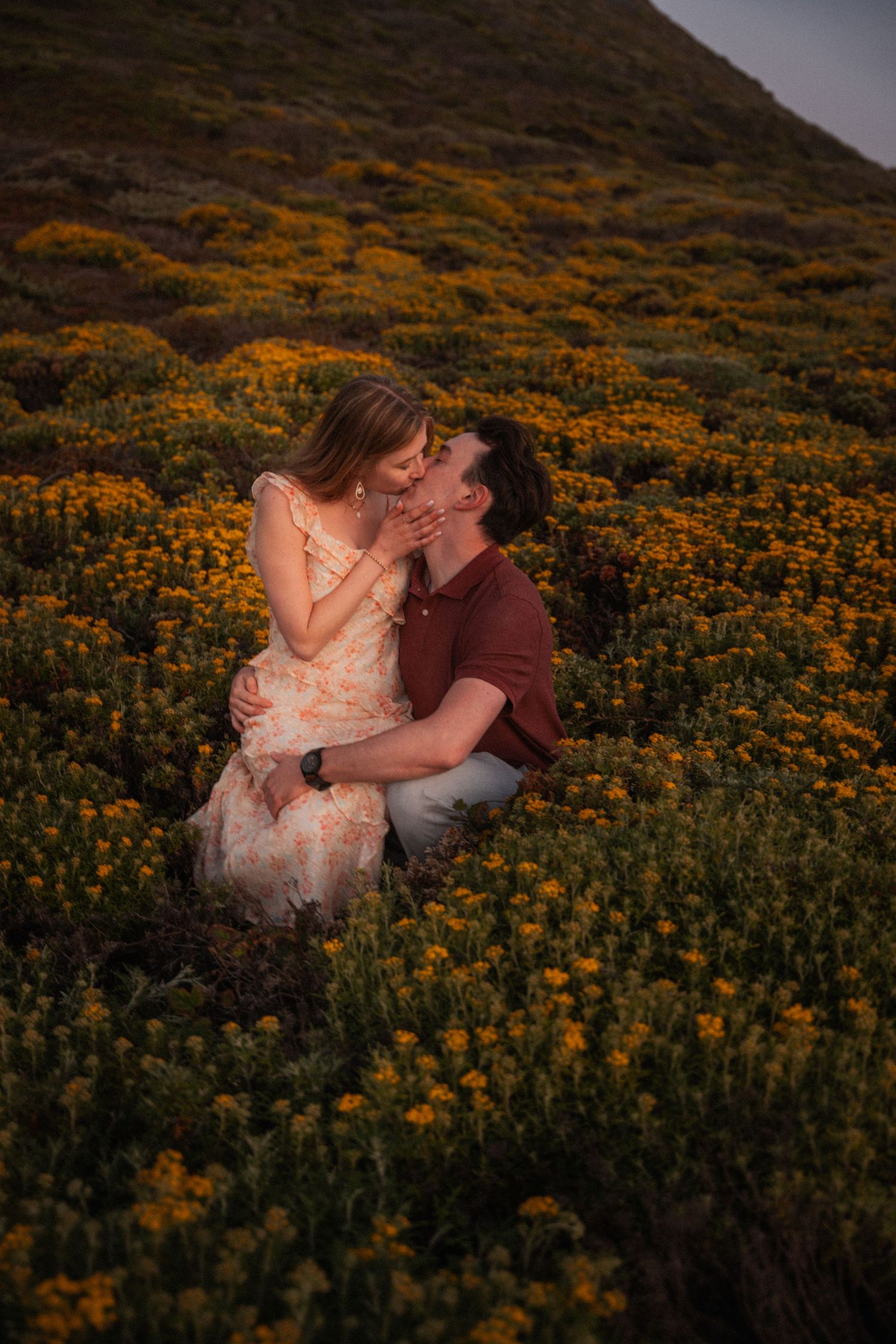 Couple kissing romantically among vibrant yellow wildflowers on a hillside during golden hour sunset.