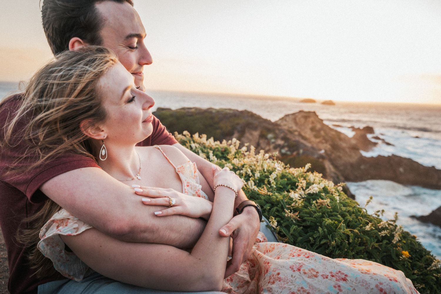 Couple embracing tenderly on rocky cliffs overlooking the ocean at sunset.