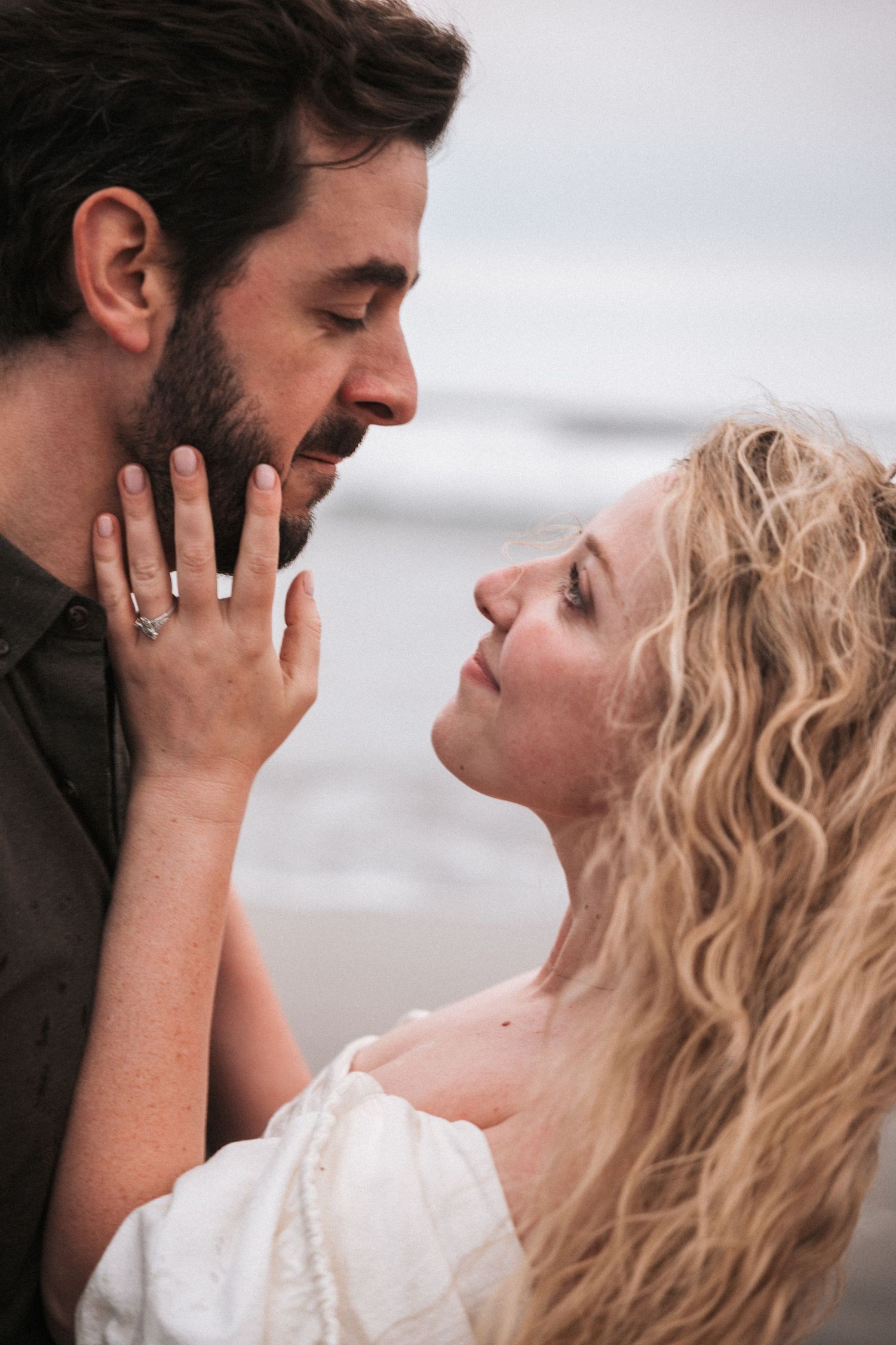 Couple sharing a romantic moment at the beach, woman with curly blonde hair touching mans bearded face, wearing engagement ring.