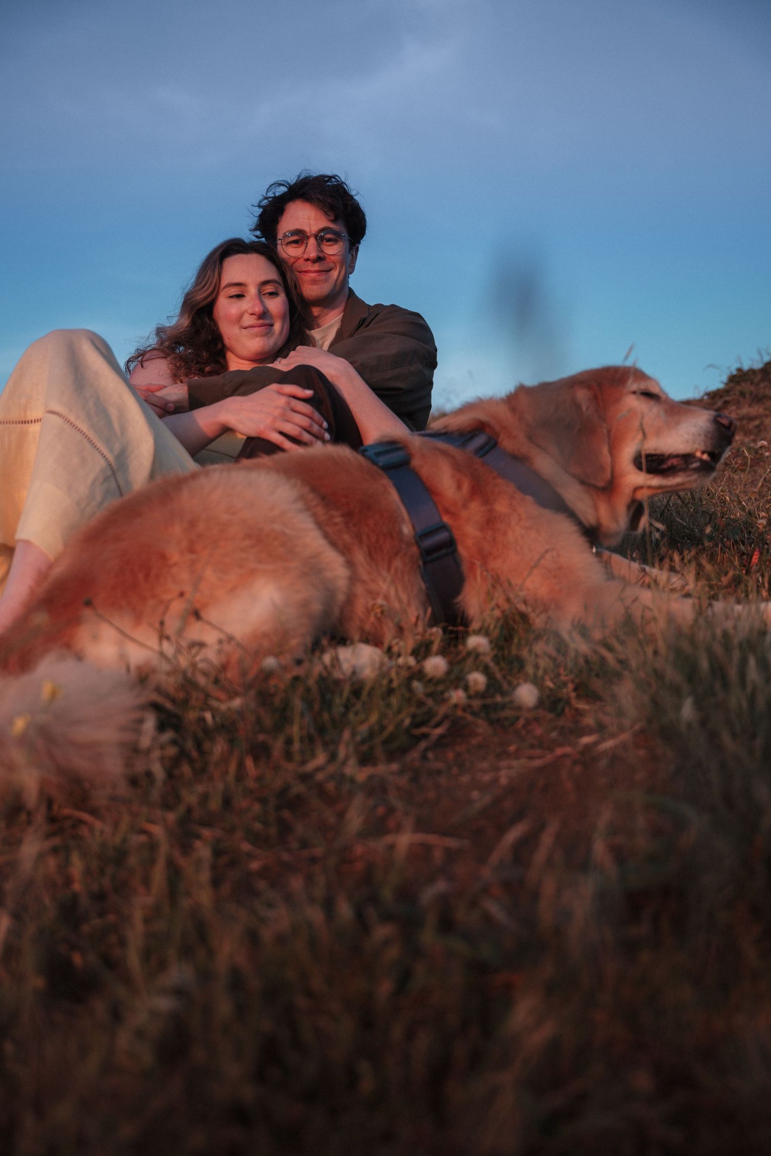 Couple sitting together on a grassy hillside at dusk with their golden retriever dog.