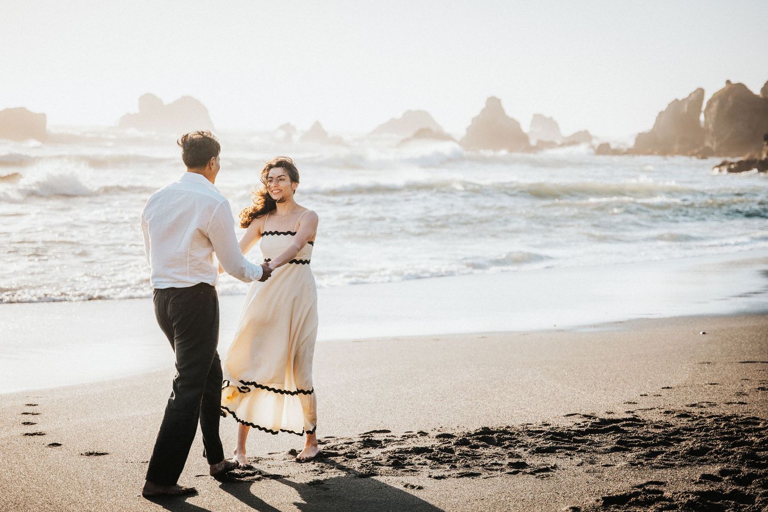 Couple dancing romantically on a sunlit beach, woman in elegant cream maxi dress holding hands with man in white shirt.