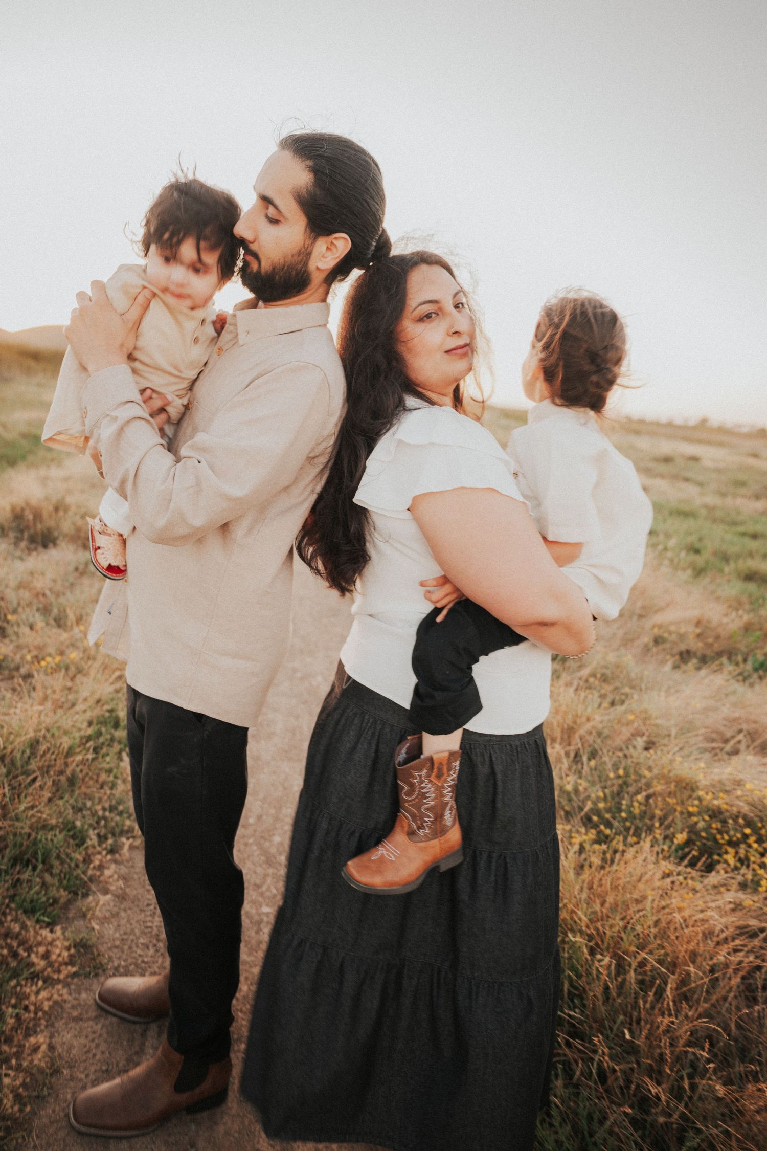 Parents standing back to back each holding a child in a golden field at sunset.