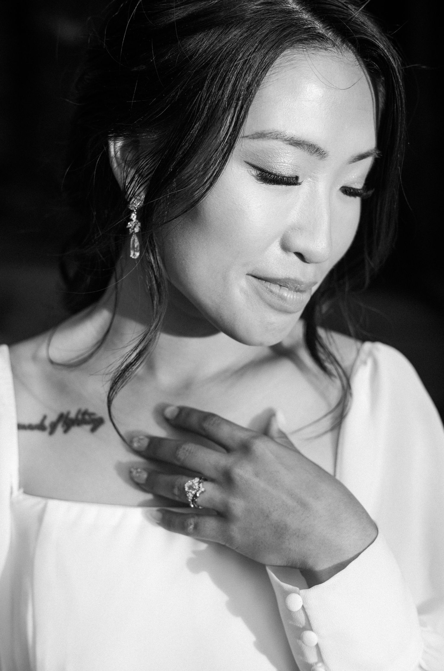 Black and white portrait of a bride in white dress with ring and tattoo, looking down gracefully.