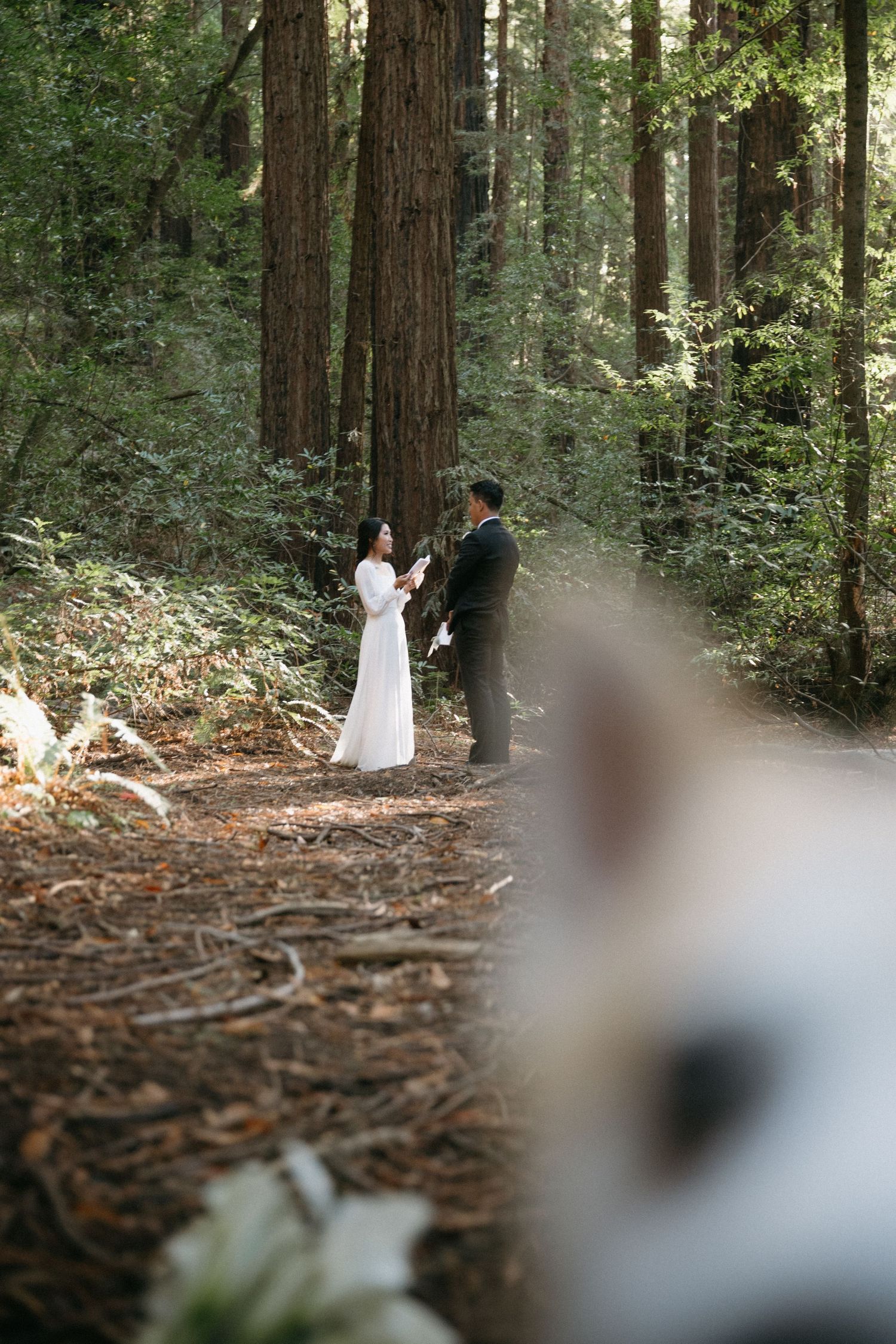 Bride and groom exchange vows on a forest path surrounded by towering redwood trees during an intimate woodland elopement.