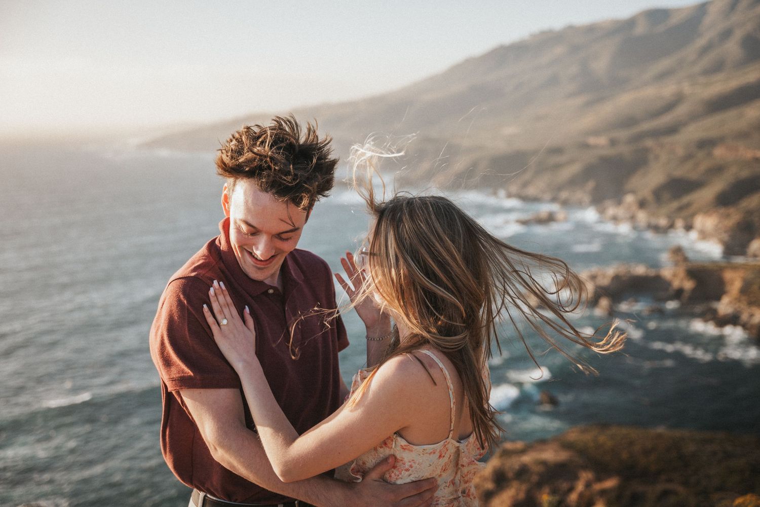 A joyful couple embraces on a coastal cliff at sunset with ocean waves and mountains in the background.