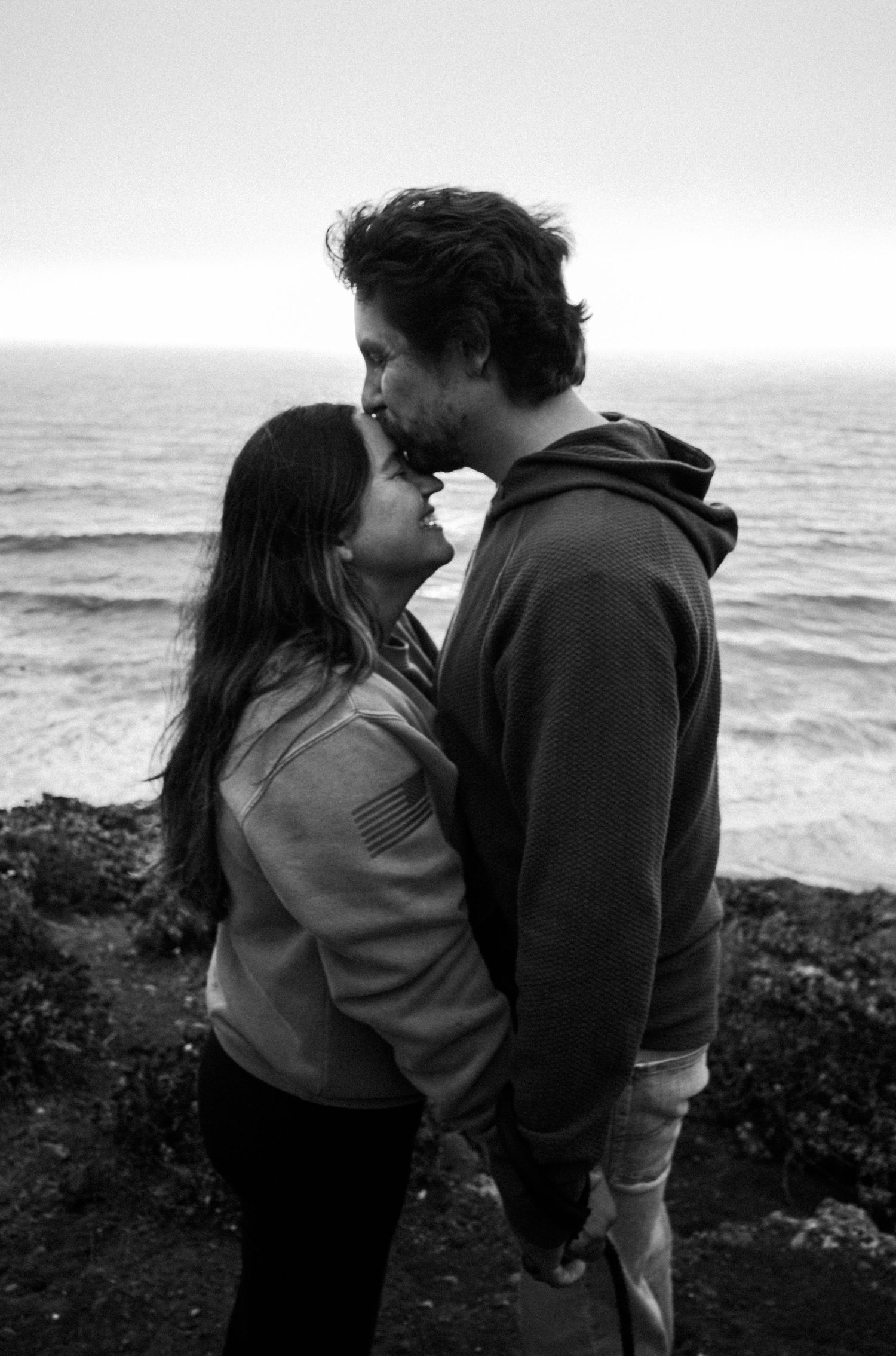 Black and white photo of a romantic couple sharing a tender moment by the ocean on a rocky coastline.