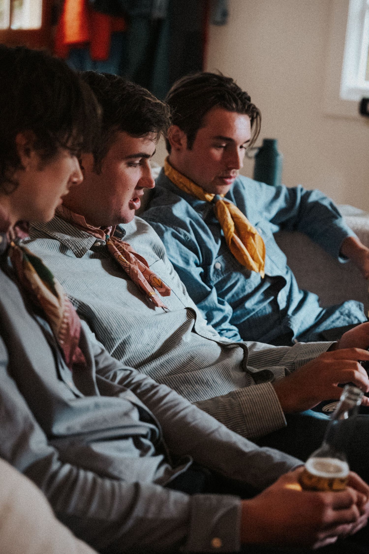 Three young men in matching blue shirts and yellow neckerchiefs sit together on a couch, one holding a drink.