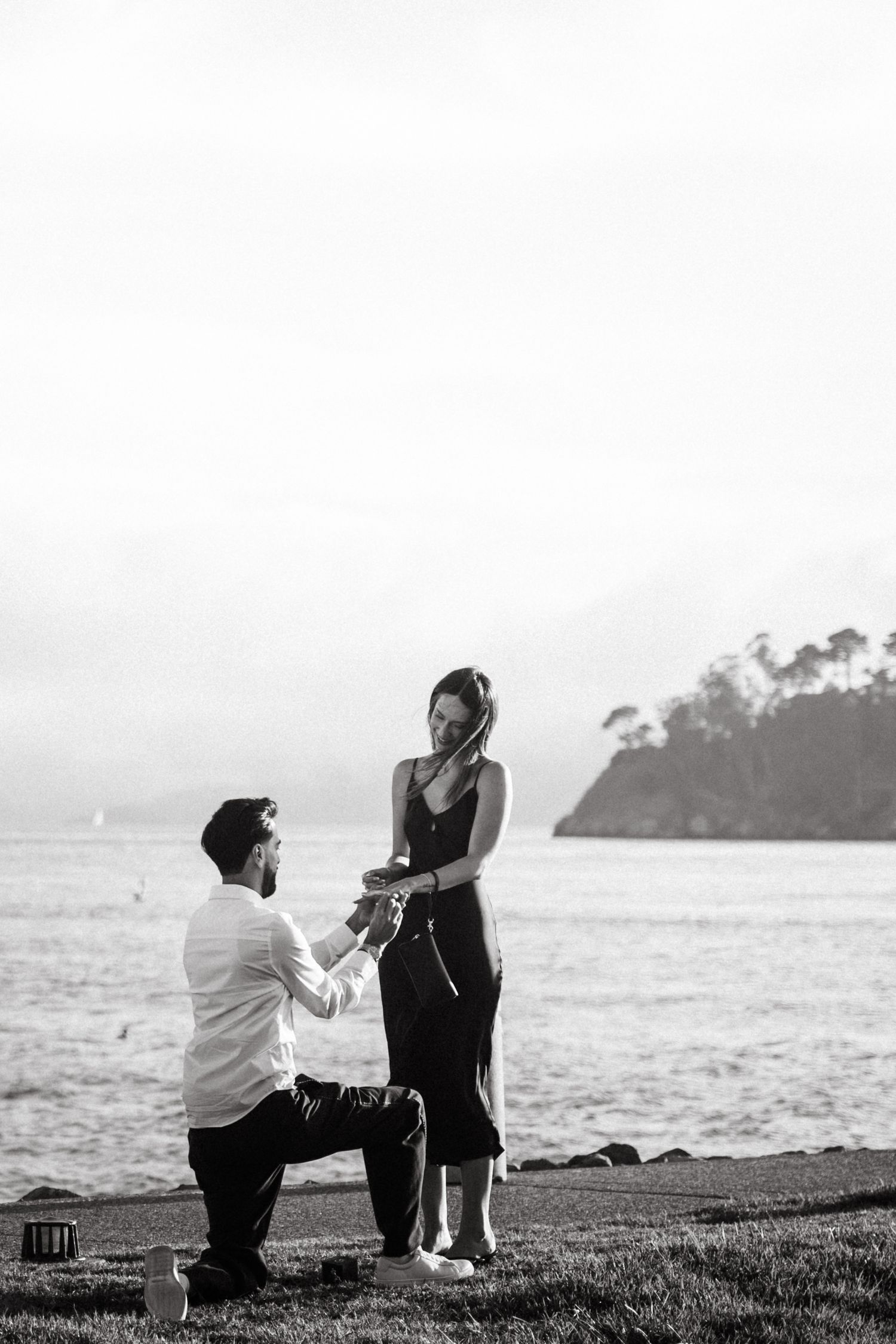 Black and white photo of a man on one knee proposing to a woman by the ocean shore.