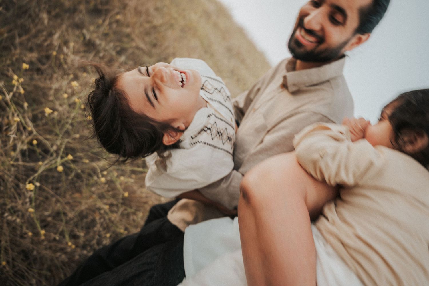 A joyful family moment with a bearded father holding a laughing child outdoors in a sunny field.