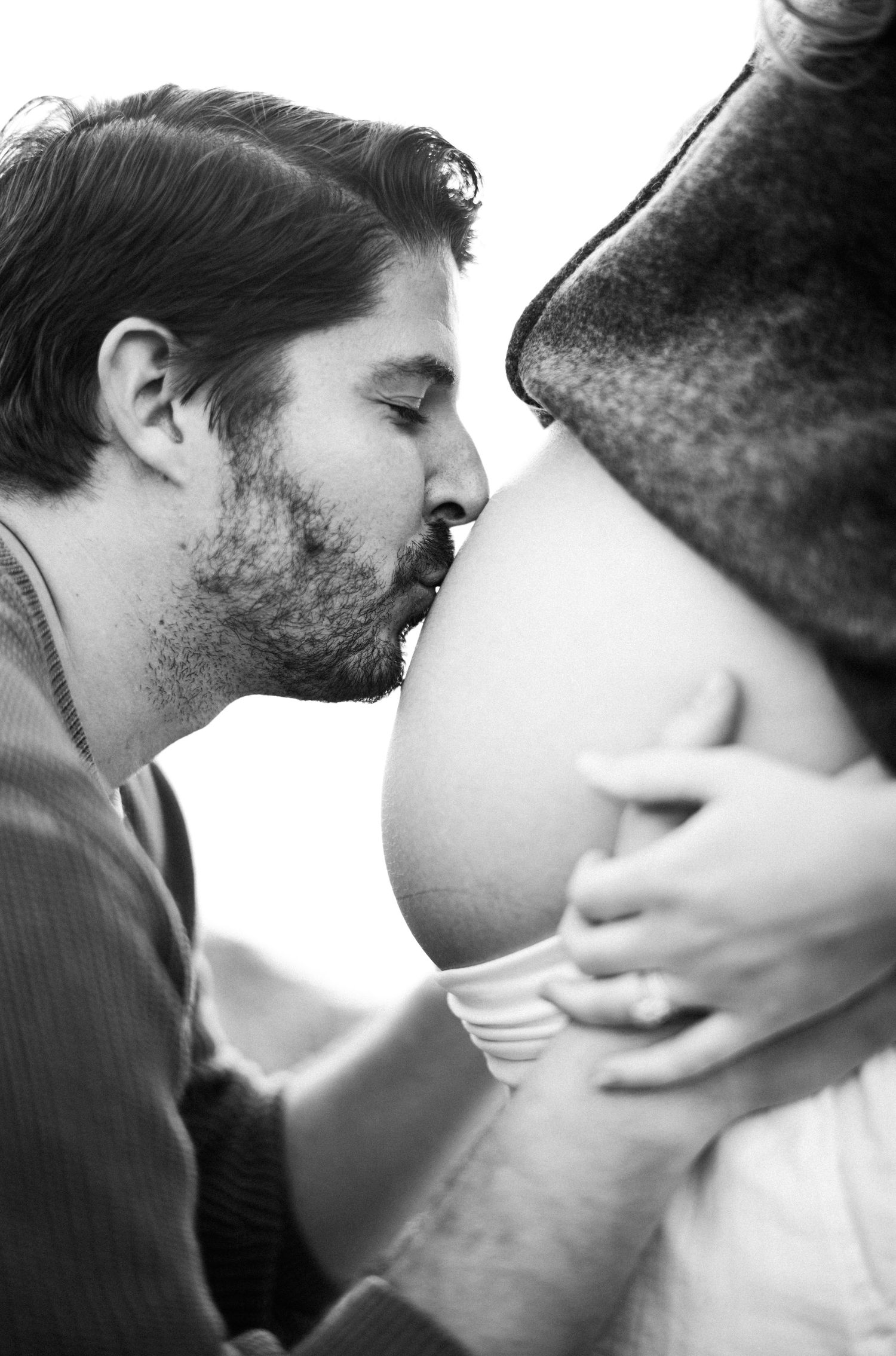 Black and white photo of a man kissing a pregnant woman's baby bump during a maternity photoshoot.