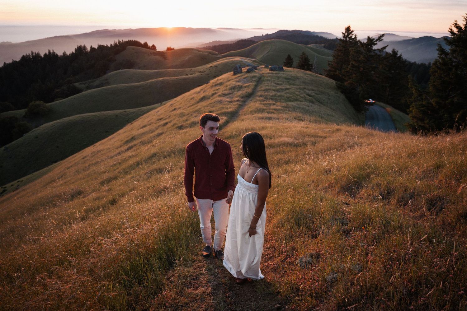 Couple walking on golden hillside trail at sunset with rolling green hills and ocean view in background.