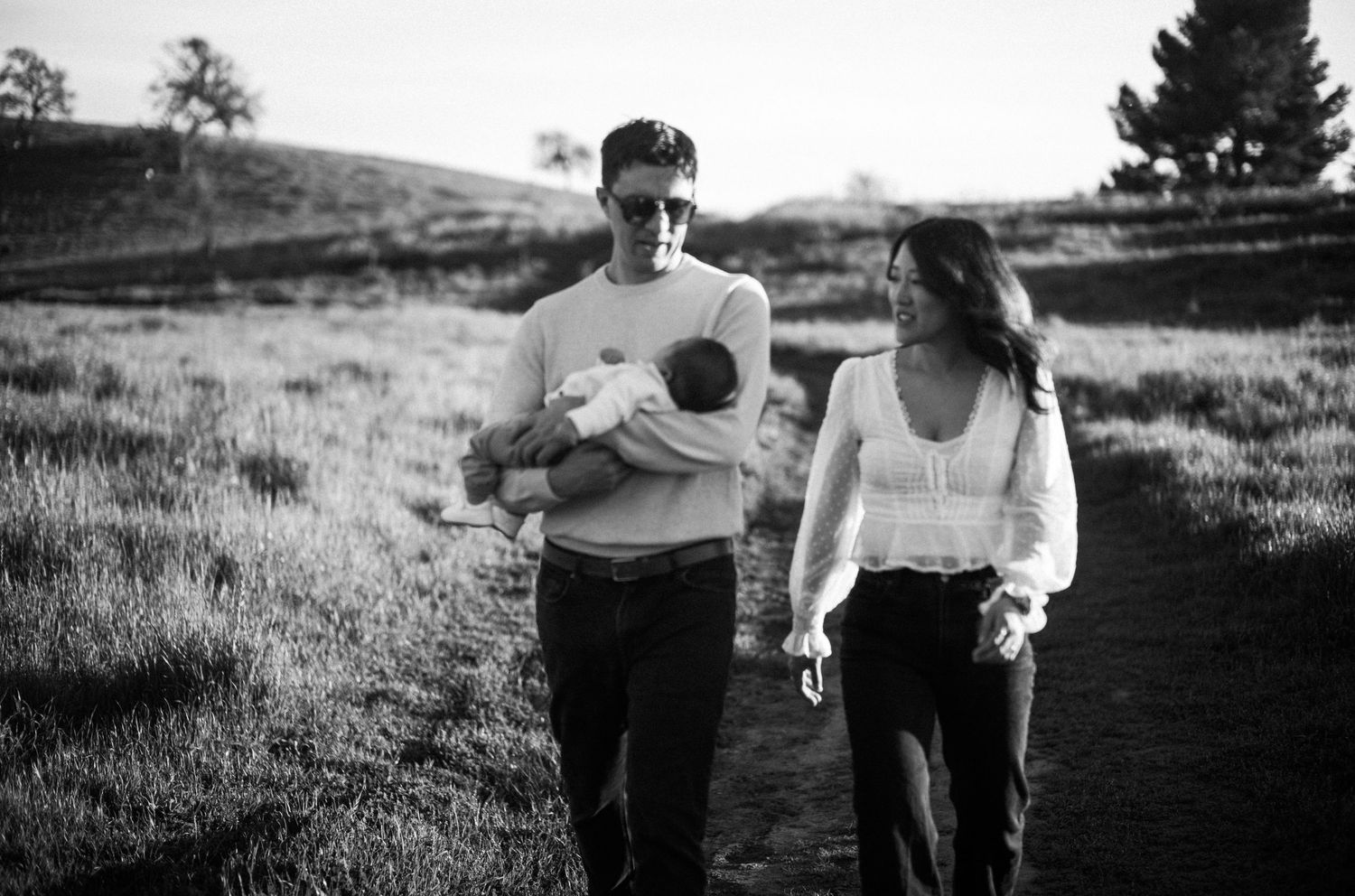 Black and white photo of a couple walking outdoors, man holding a newborn baby, woman in white top, grassy hills behind.