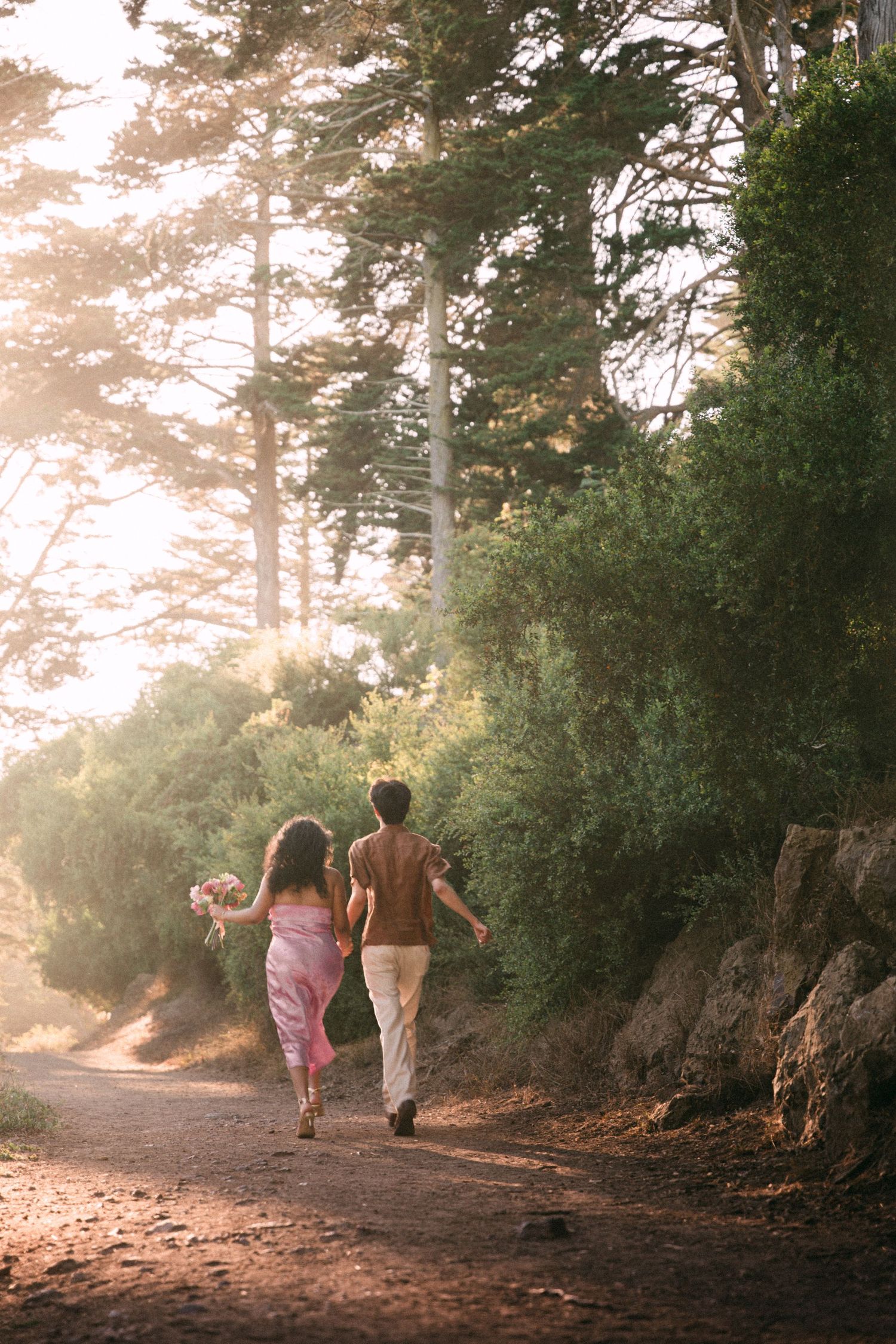 Couple walking hand-in-hand down a misty forest path, woman in pink dress holding flowers, bathed in golden sunlight.