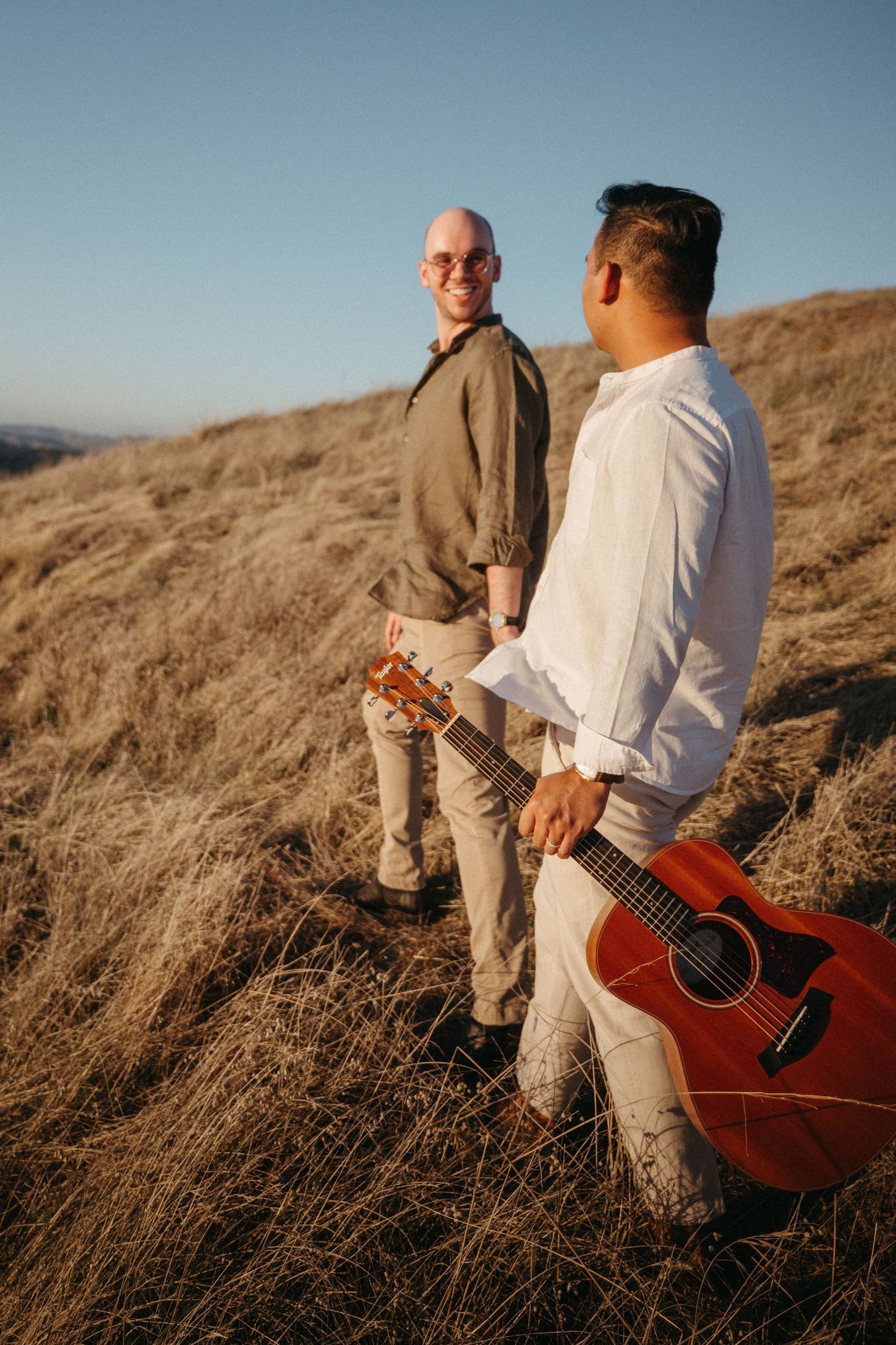 Two men stand on a golden hillside at sunset, one holding an acoustic guitar, enjoying a scenic outdoor moment.