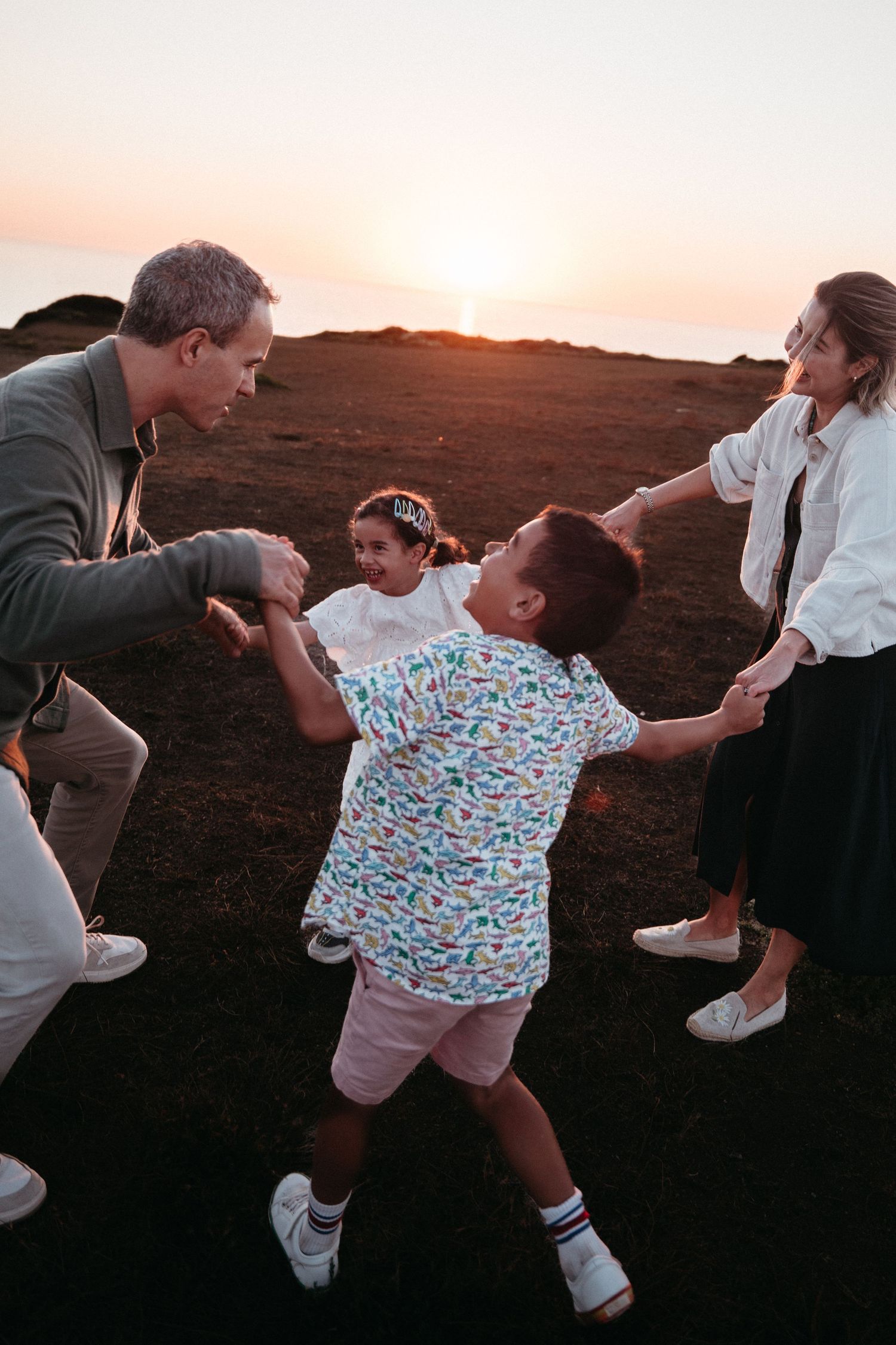 Family of four joyfully holding hands in a circle at sunset on a rocky landscape.