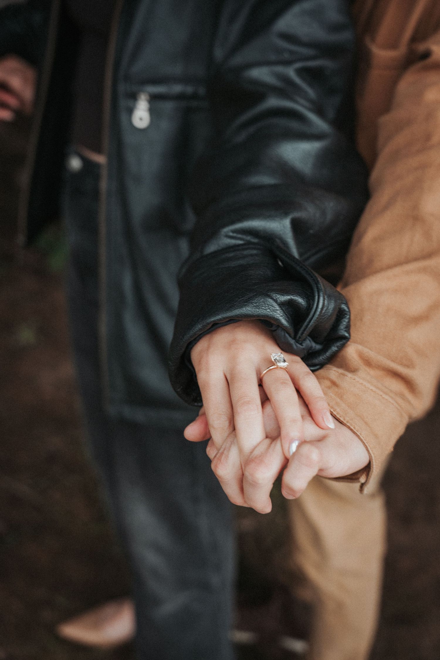 Two people holding hands with an engagement ring visible, wearing leather and tan jackets.