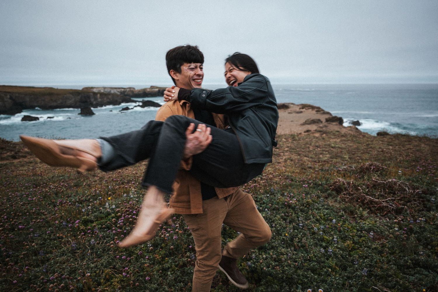 Couple laughing joyfully on a coastal cliff, man carrying woman in his arms with ocean waves in background.
