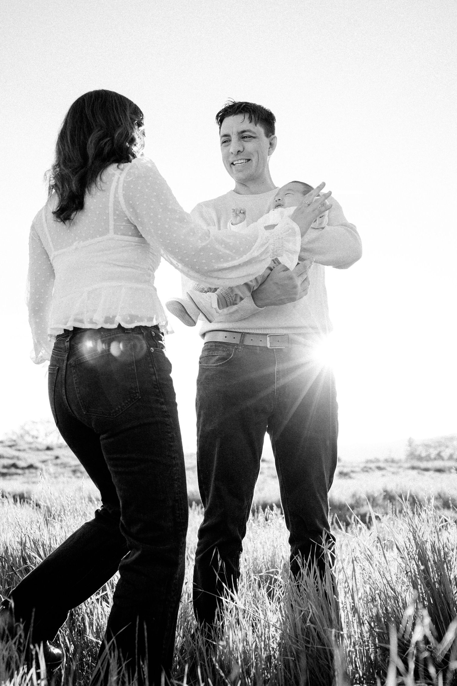 Couple with newborn baby standing in golden sunlit grass field black and white portrait.