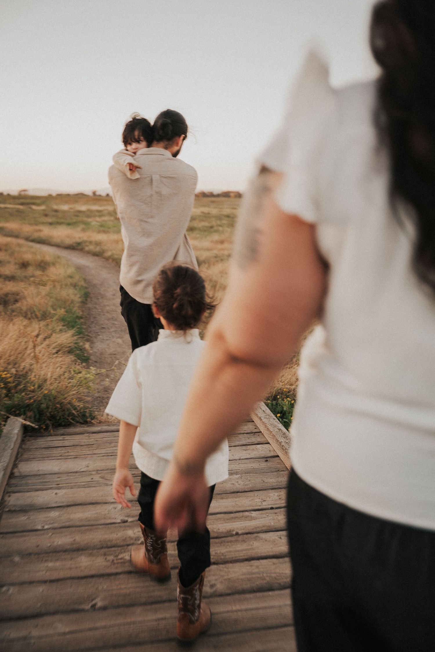 Father carrying child on wooden boardwalk path through golden fields at sunset with family following.